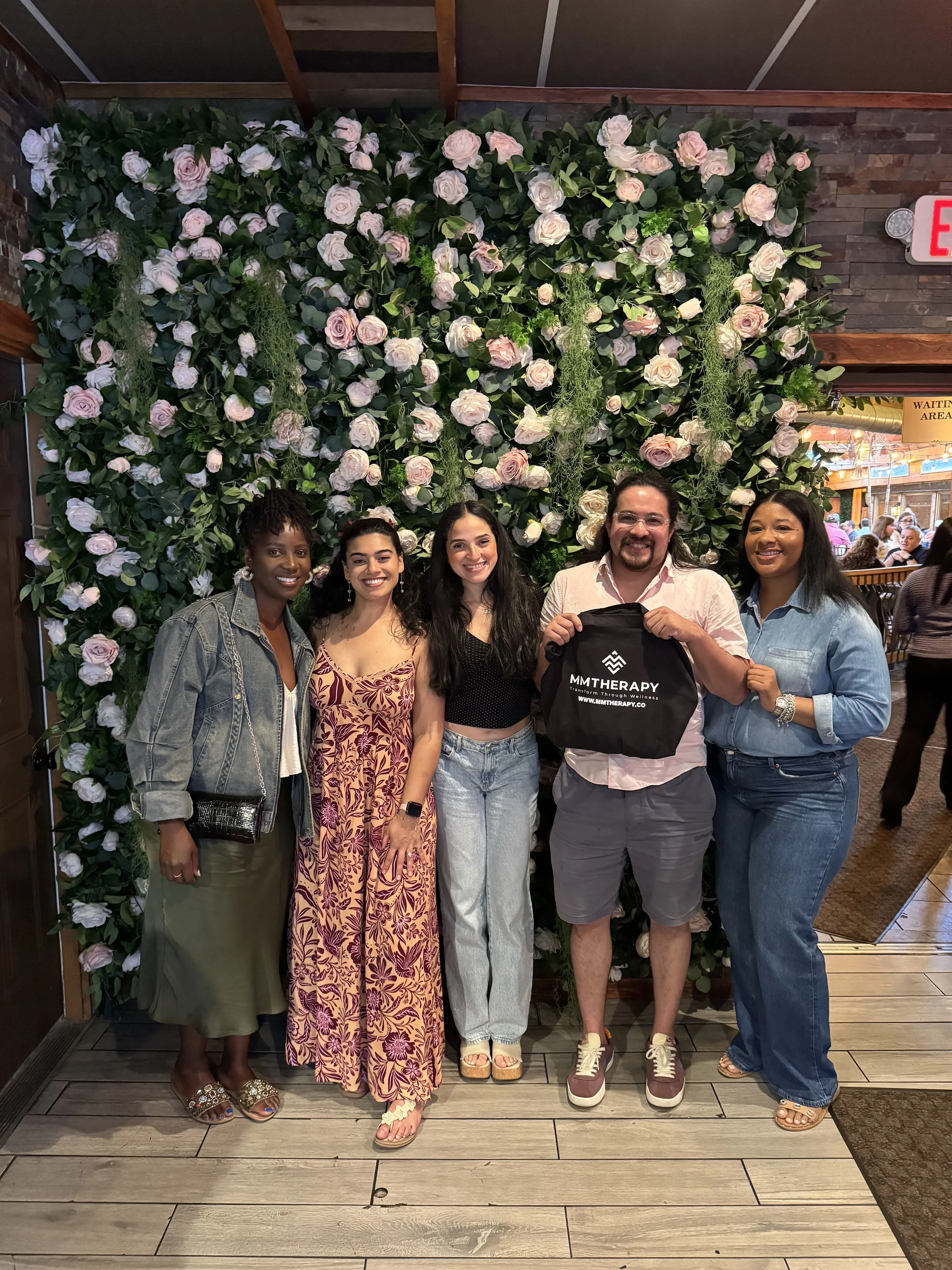 Group of five people standing in front of a floral wall with pink and white roses inside a restaurant or café. They are smiling, with the man in the center holding a black bag with white text that reads 'MMTHERAPY'.