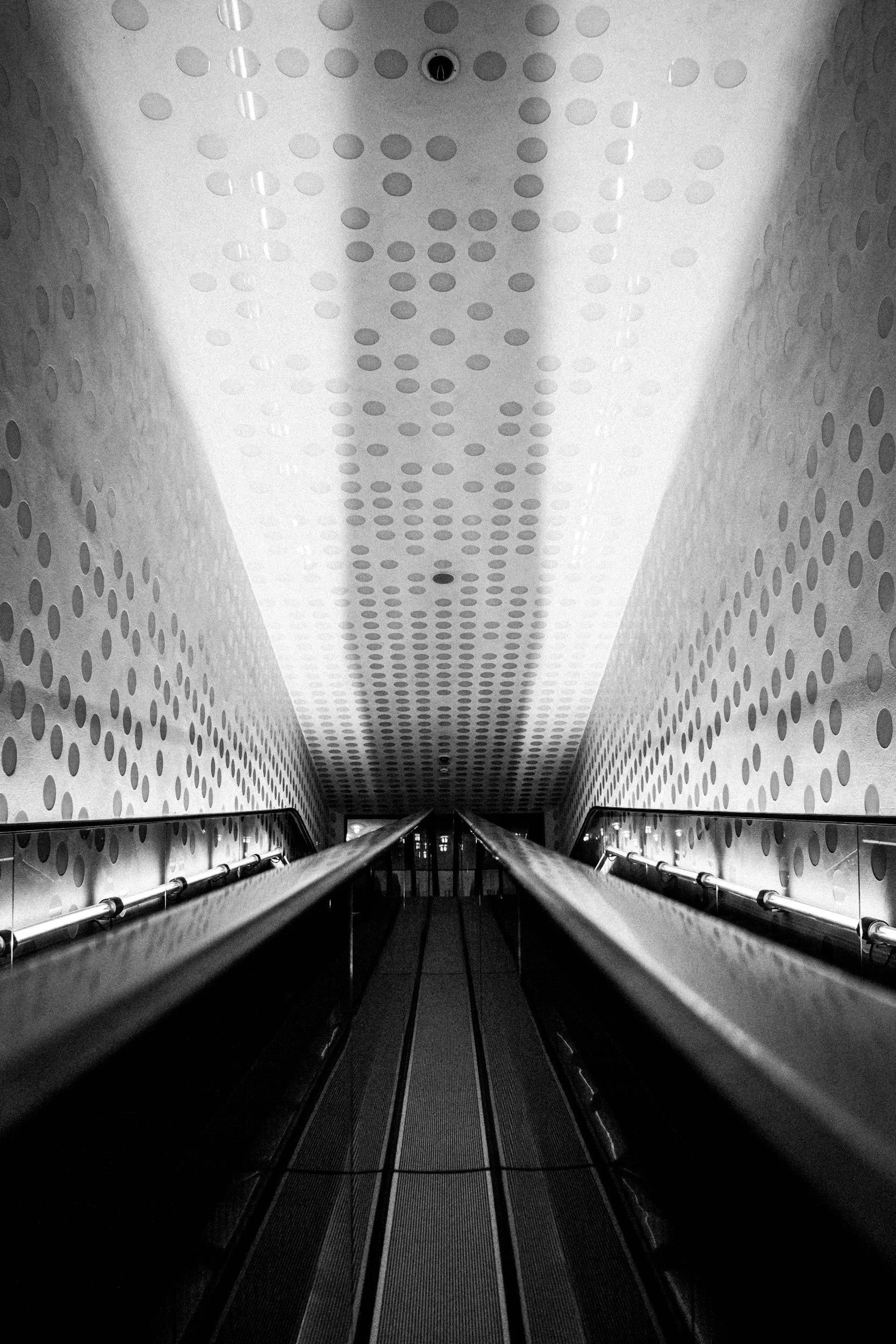 Black and white photo of an escalator in a modern building with dotted pattern walls and ceiling.
