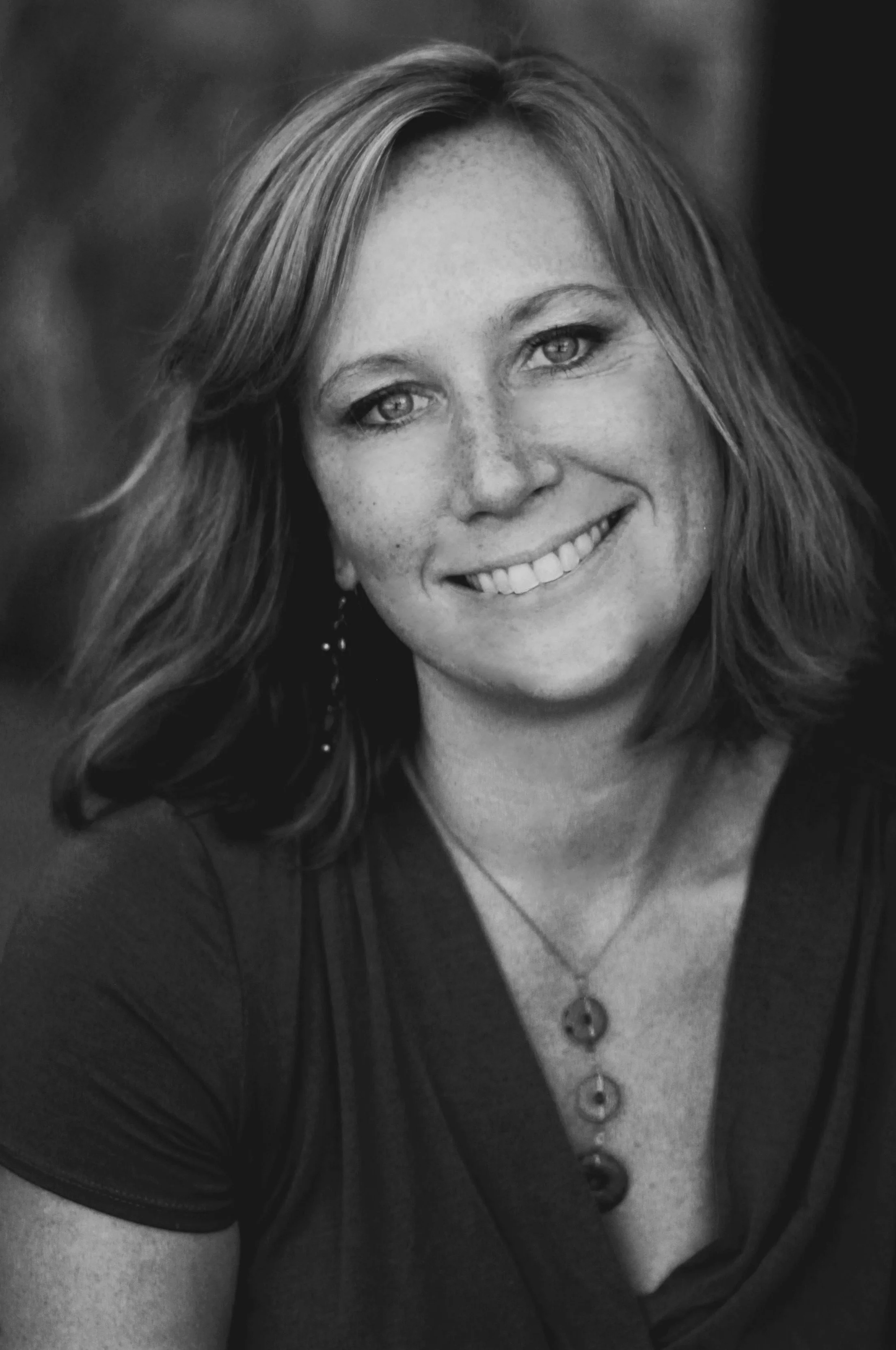 Black and white portrait of a woman smiling, with shoulder-length hair, wearing a necklace with three pendants and earrings.