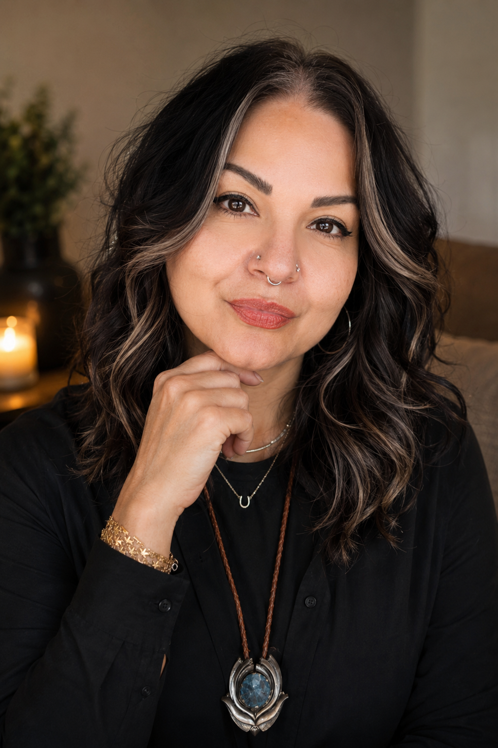 A woman with wavy, dark brown hair with gray highlights, wearing a black shirt, jewelry including necklaces, a bracelet, and a nose ring, posing indoors with a thoughtful expression, resting her chin on her hand, with a candle and potted plant in the background.