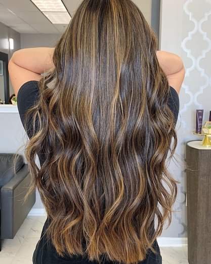 Back view of a woman with long, wavy, light brown hair with blonde highlights in a salon setting.