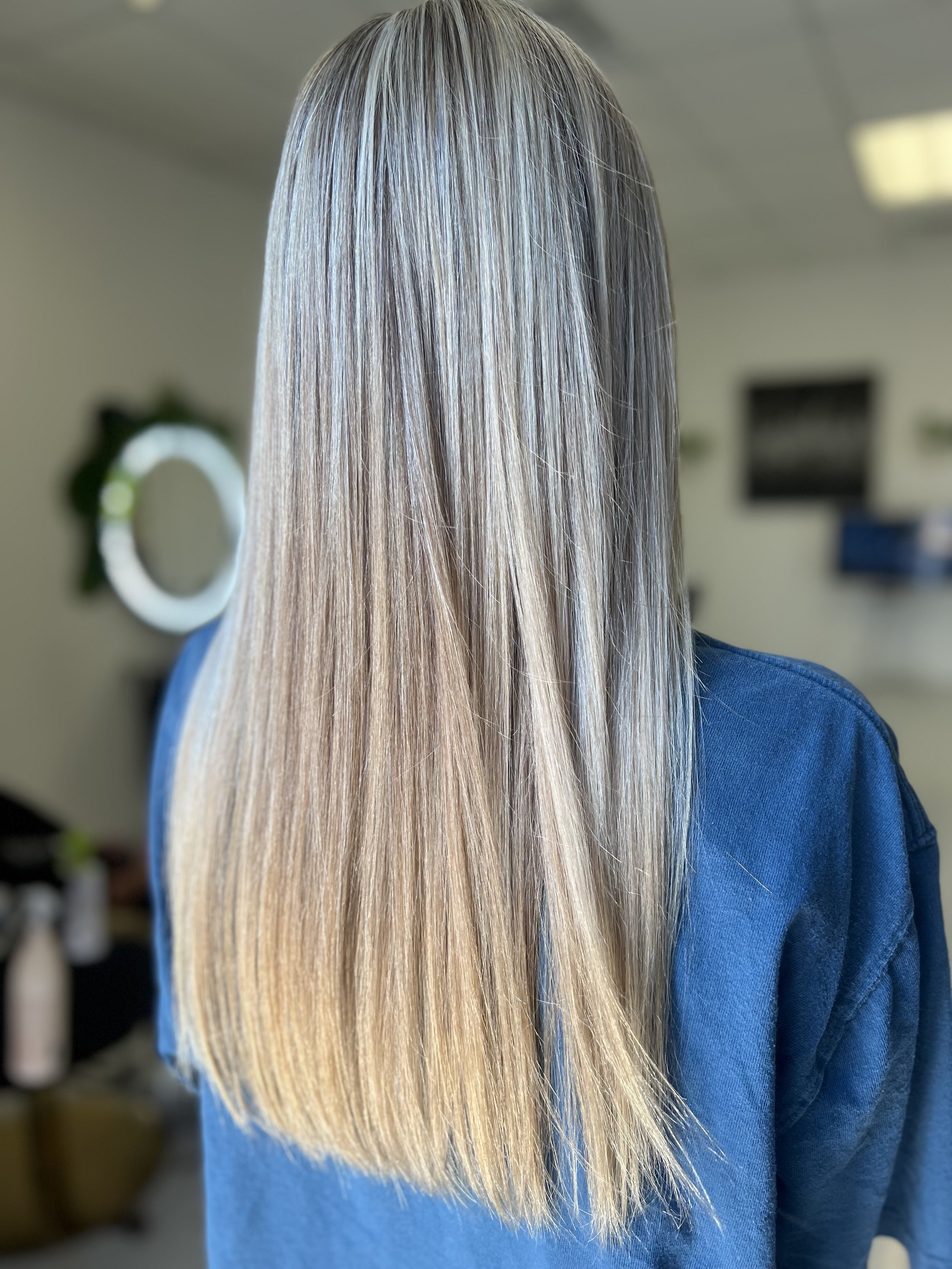 Back view of a woman with long, straight, blonde hair wearing a blue top, in a salon setting.