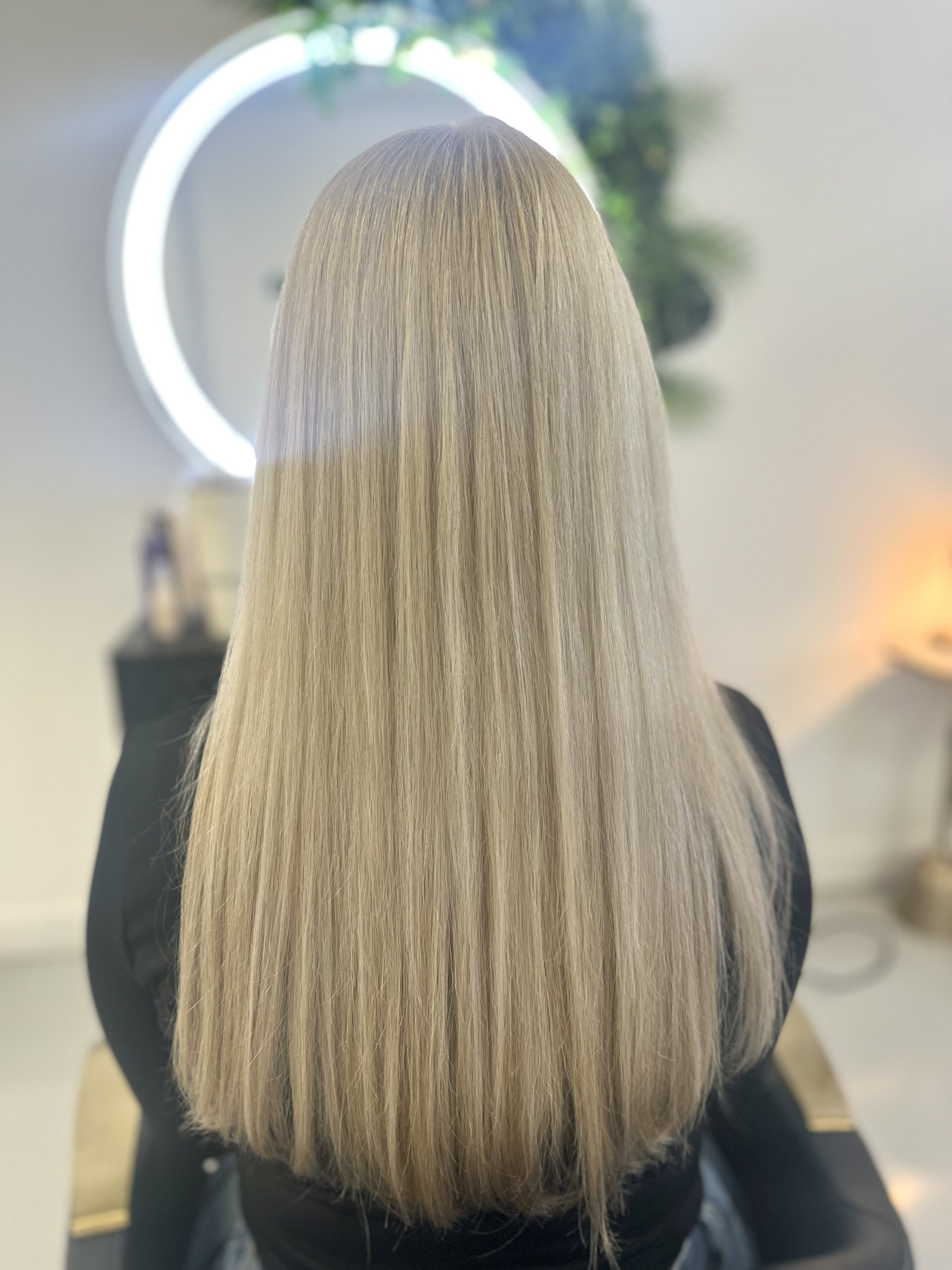 Back of a person with long, straight, blonde hair sitting in a salon, with a mirror and hair styling tools in the background.