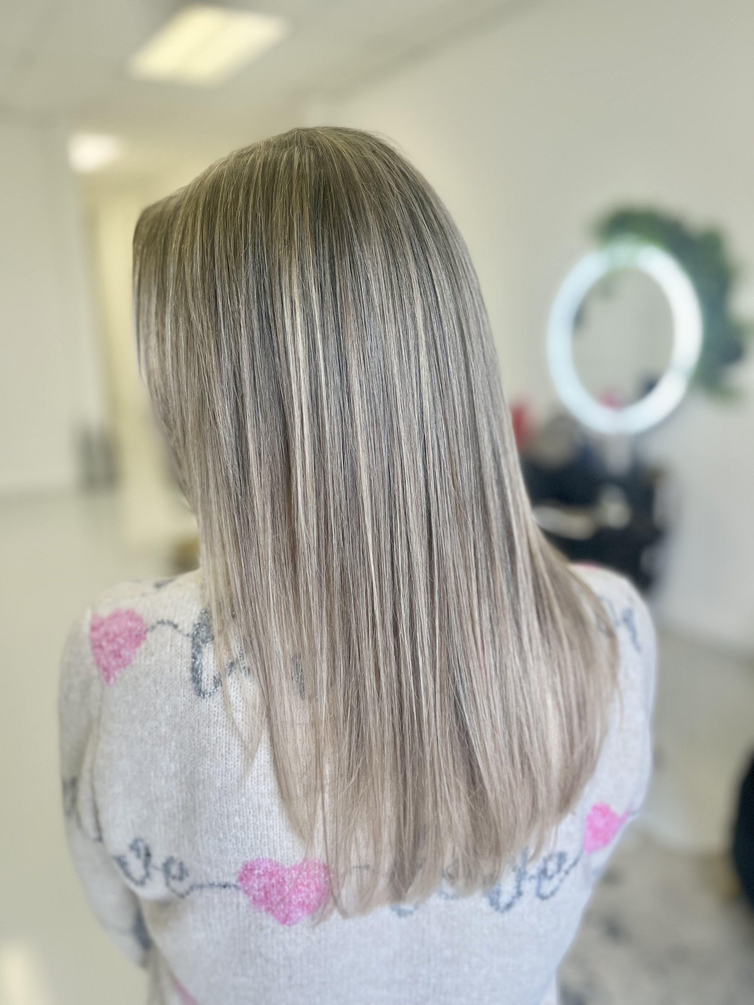 Back view of a woman with straight, shoulder-length blonde hair, wearing a white sweater with pink hearts and black cursive writing, in a salon with a ring light visible in the background.