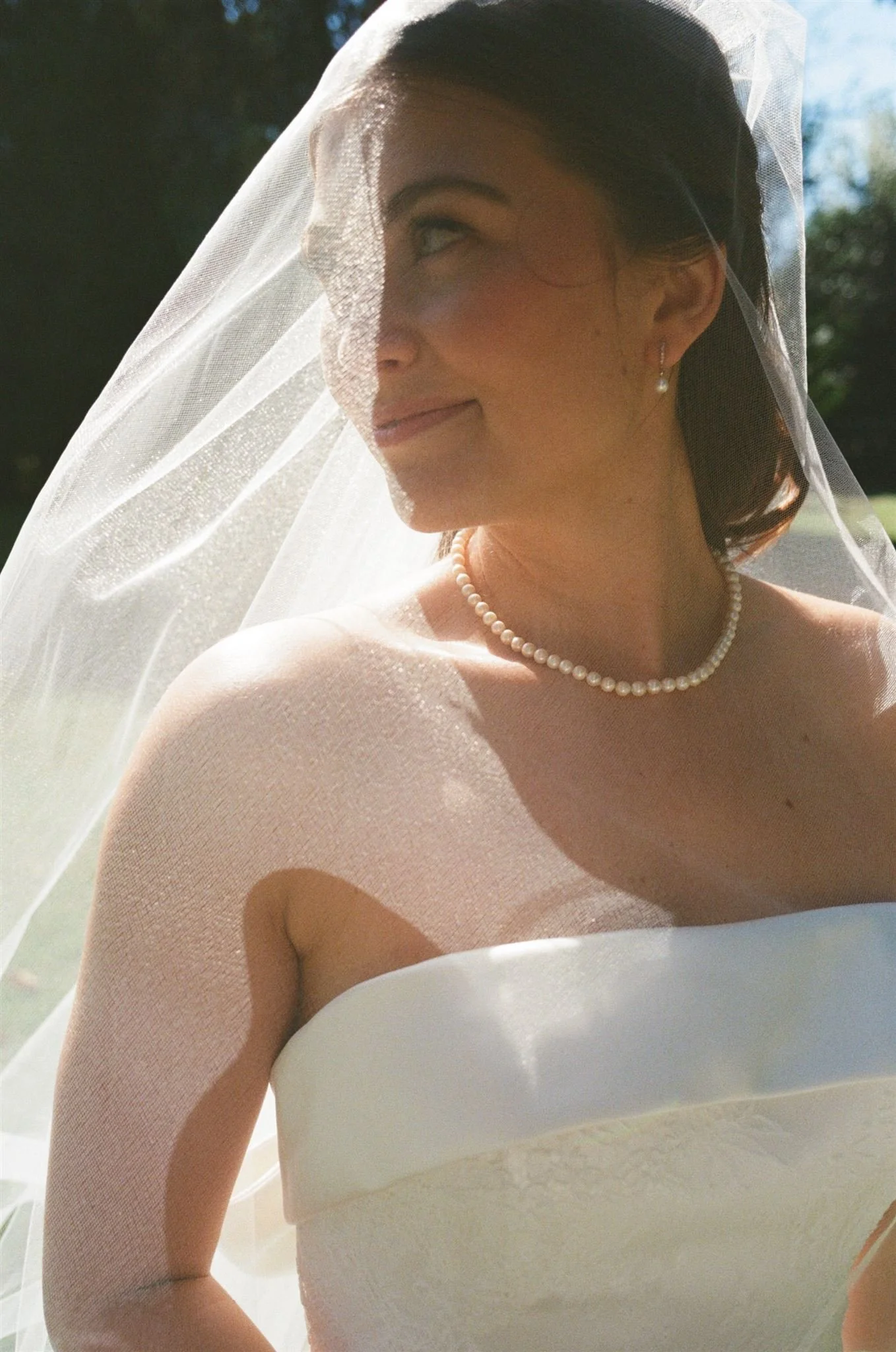 A bride in a white strapless wedding dress wearing a pearl necklace and earrings, with a sheer veil covering her face, smiling outdoors on a sunny day.