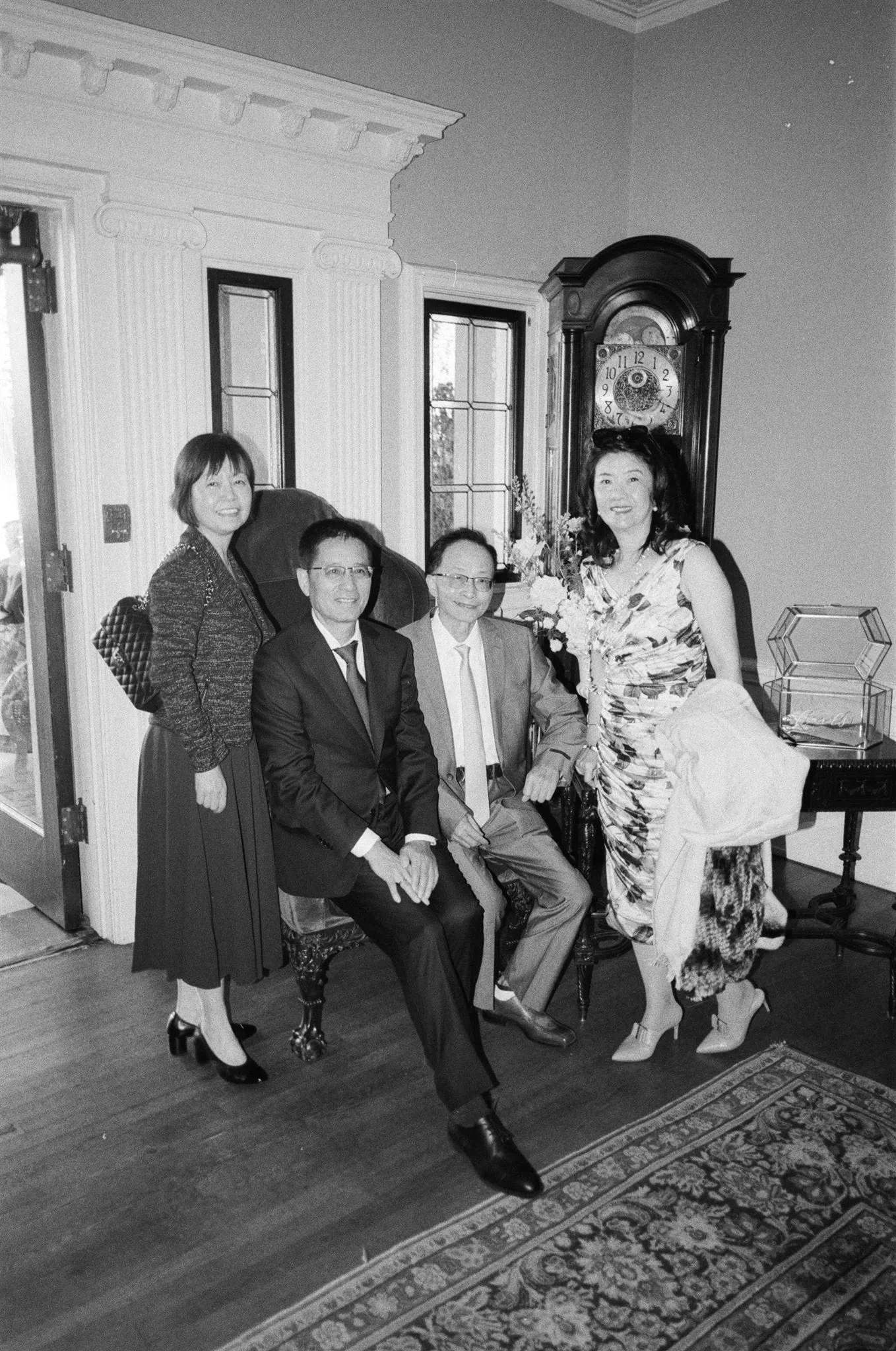Black and white photo of five people in a room with antique furniture and a grandfather clock, posing for a picture. Two women stand and three men sit, dressed formally, smiling at the camera during a wedding.