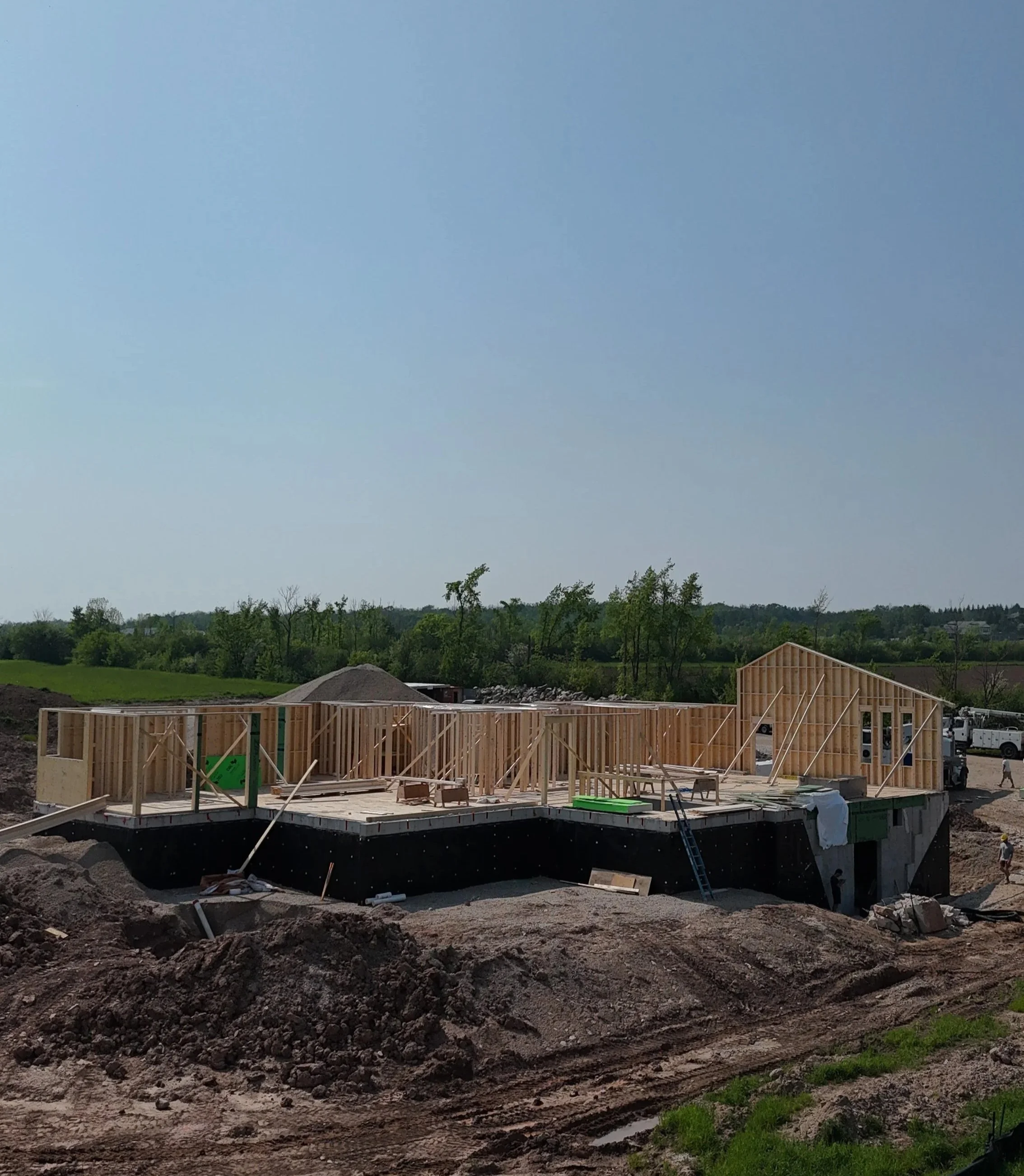 Construction site with a partially built wooden house frame on a foundation, surrounded by dirt and construction materials, with a clear blue sky and green trees in the background.