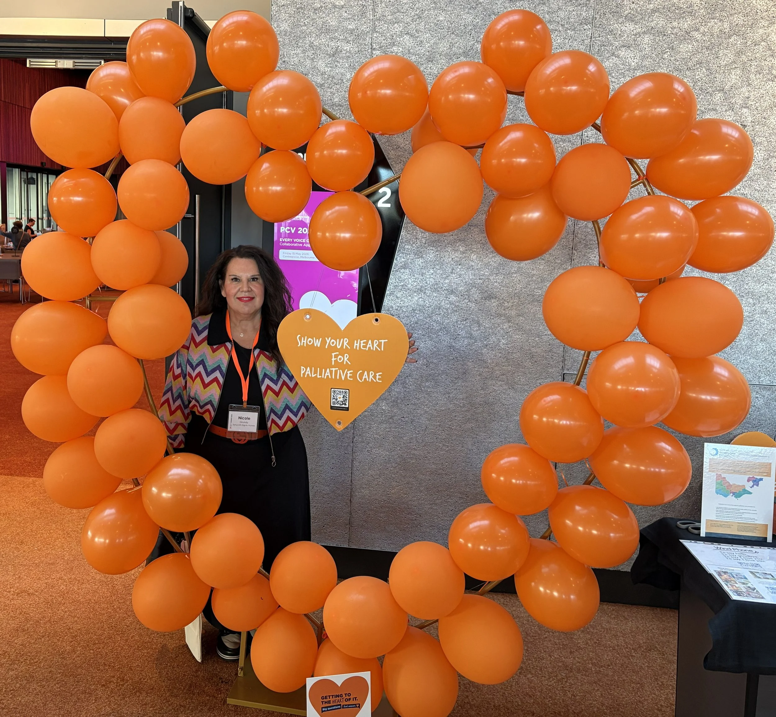 A woman stands in front of an orange balloon display holding a heart-shaped sign that reads, 'Show your heart for palliative care.' The display consists of numerous orange balloons arranged in a circular pattern. There are informational posters and a