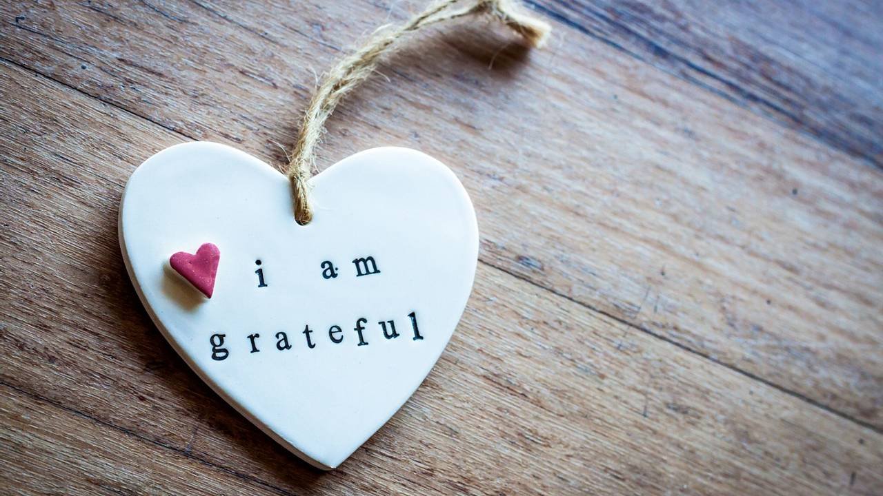 White heart-shaped ornament with a small pink heart and the words "i am grateful" printed on it, resting on a wooden surface.