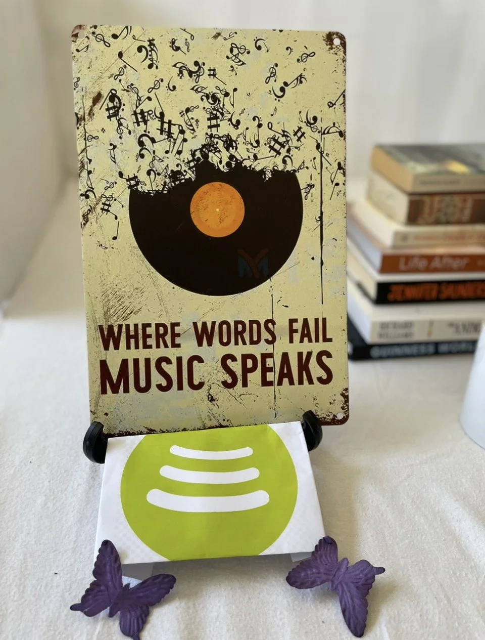 A metal sign with musical notes and symbols, featuring a black record with an orange center, and the phrase 'WHERE WORDS FAIL MUSIC SPEAKS' written on it. The sign is displayed on a stand, with small purple butterfly figurines at the base, on a white