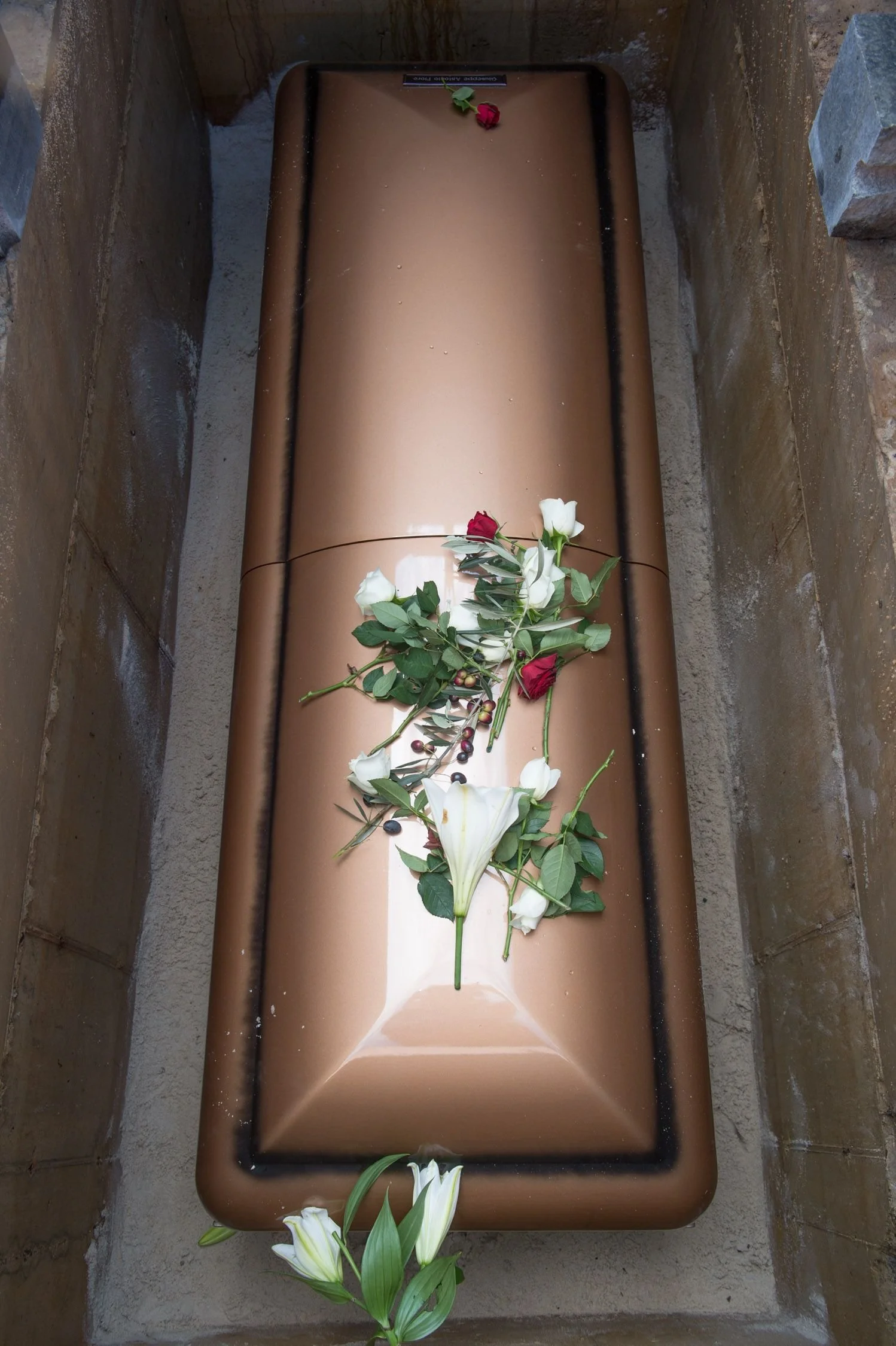 A bronze-colored coffin with flowers placed on top, inside a stone coffin or niche.