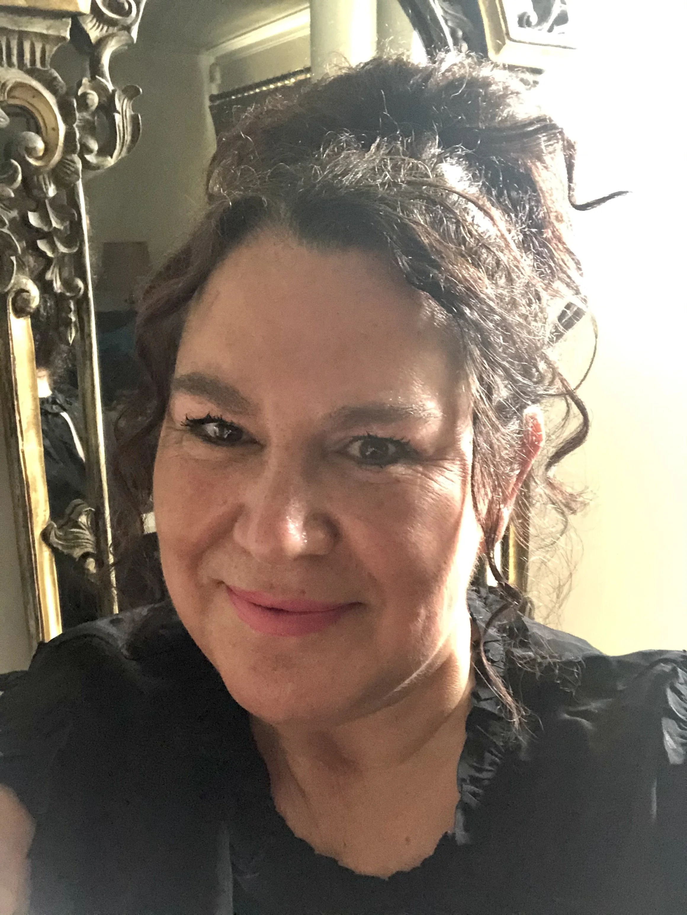 Nicole Grundy celebrant, A woman with curly hair taking a selfie indoors, with a decorative mirror and a lamp in the background.