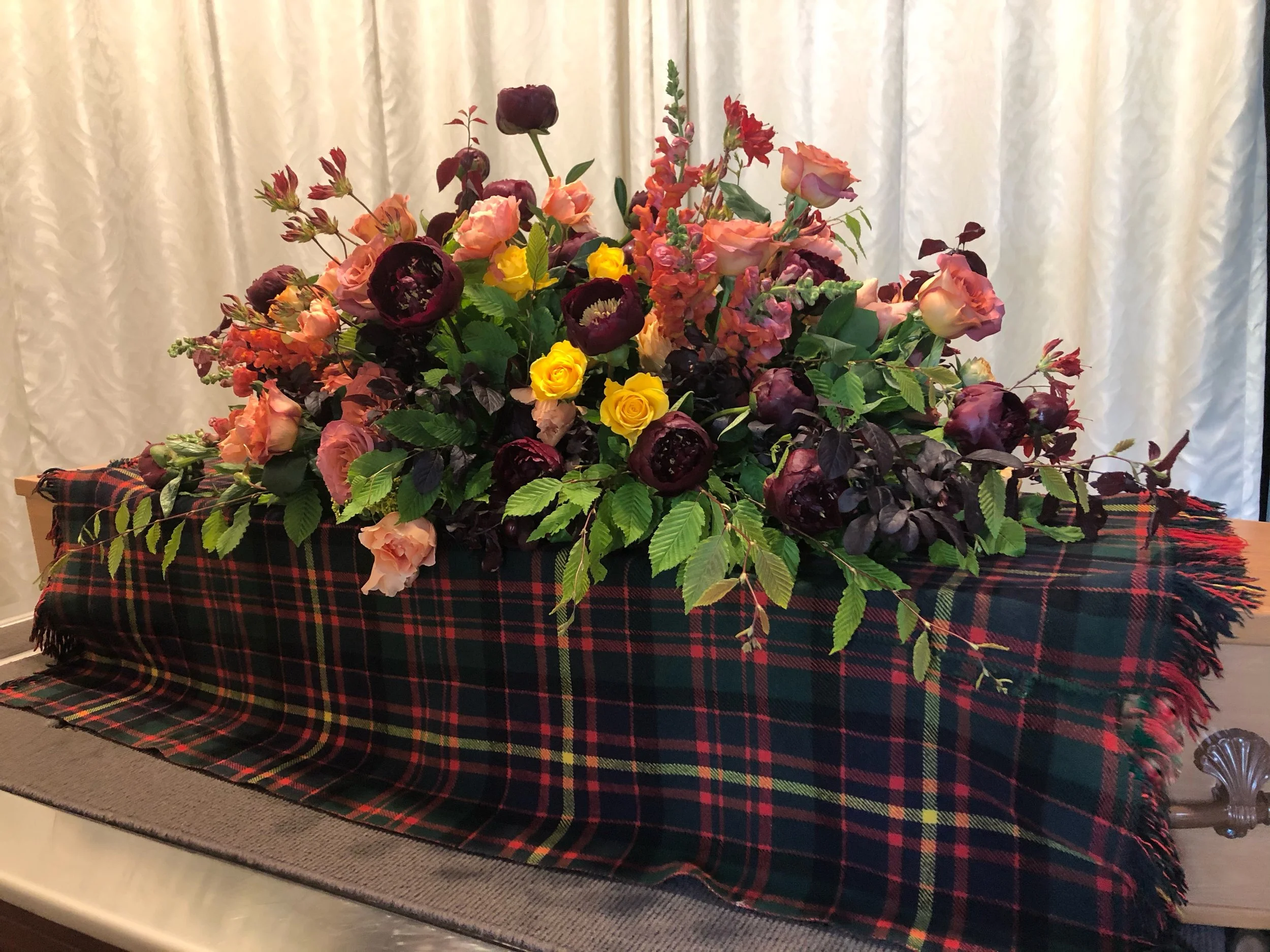 A large floral arrangement of roses, peonies, and other flowers in shades of pink, yellow, and dark purple, placed on a table with a plaid cloth in green, red, and black, with a white curtain in the background.
