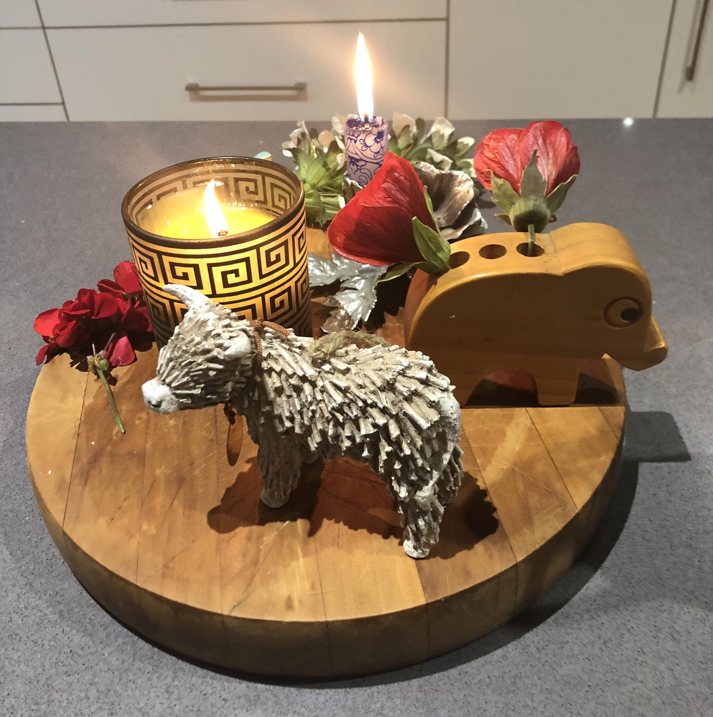 A decorative candle with a flame, surrounded by flowers and pinecones on a wooden table.