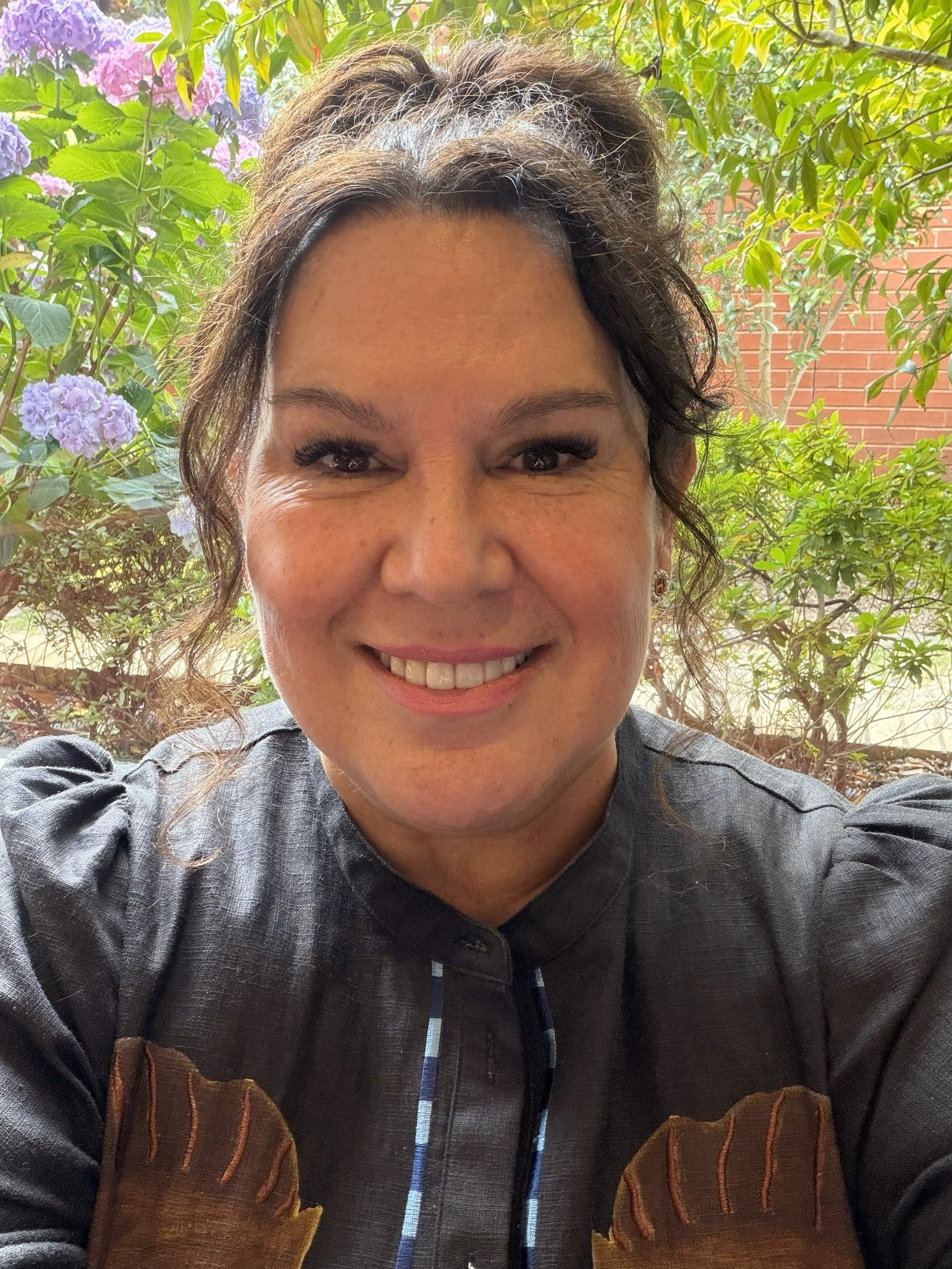 Close-up of a woman smiling outdoors with green foliage and purple flowers in the background.