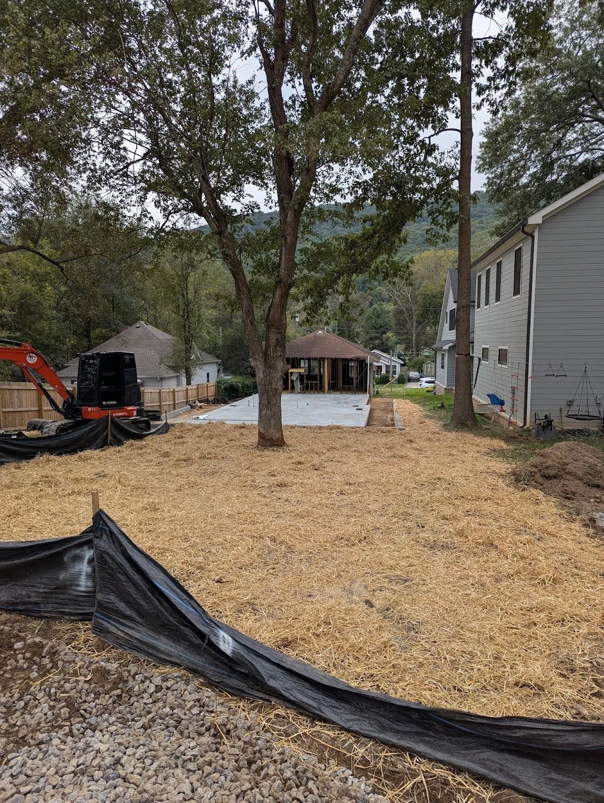 Backyard under construction with a large tree in the center, a concrete slab, a small shed, and construction equipment, with neighboring houses and a mountain in the background.