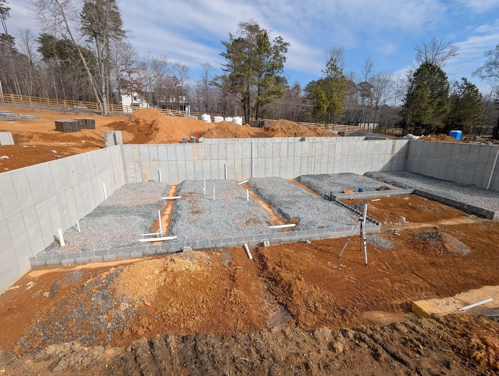 Construction site with foundation work in progress, gravel and concrete block walls, pipes, and soil around.