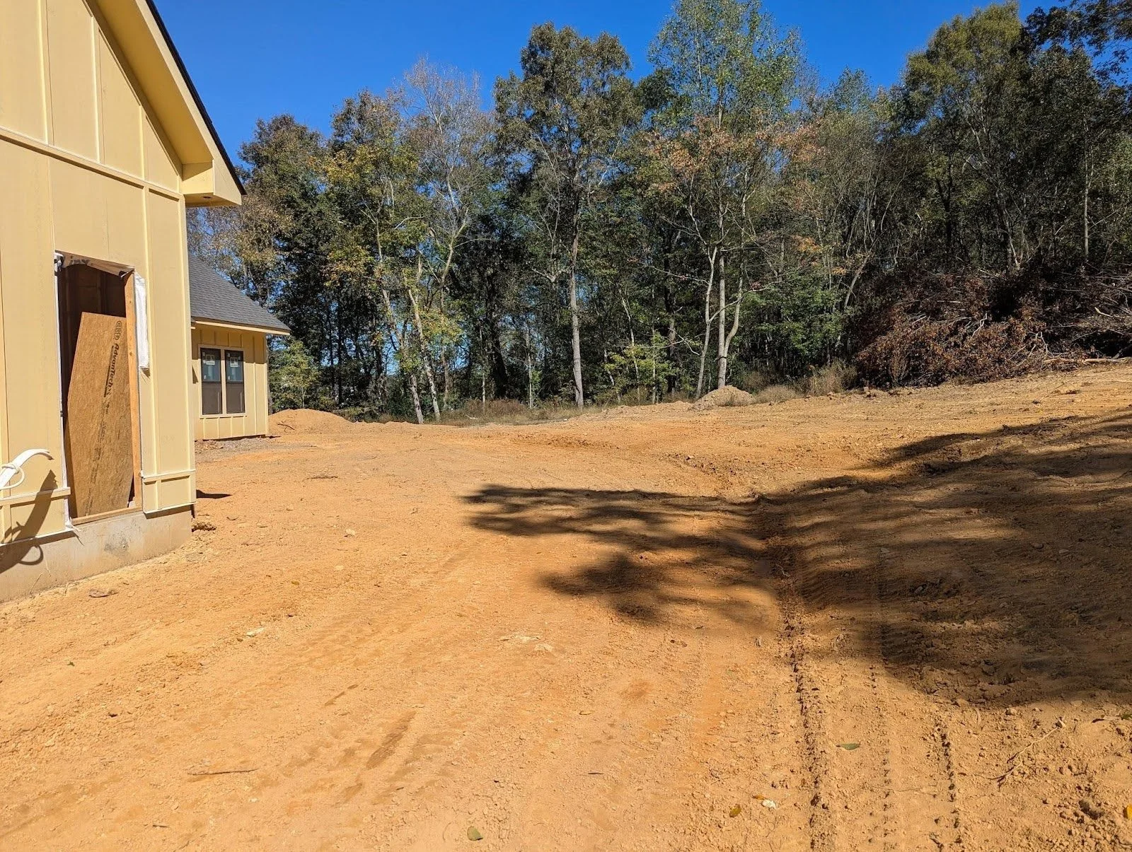 Construction site with a partially built yellow house on the left and dirt ground, surrounded by trees under a clear blue sky.