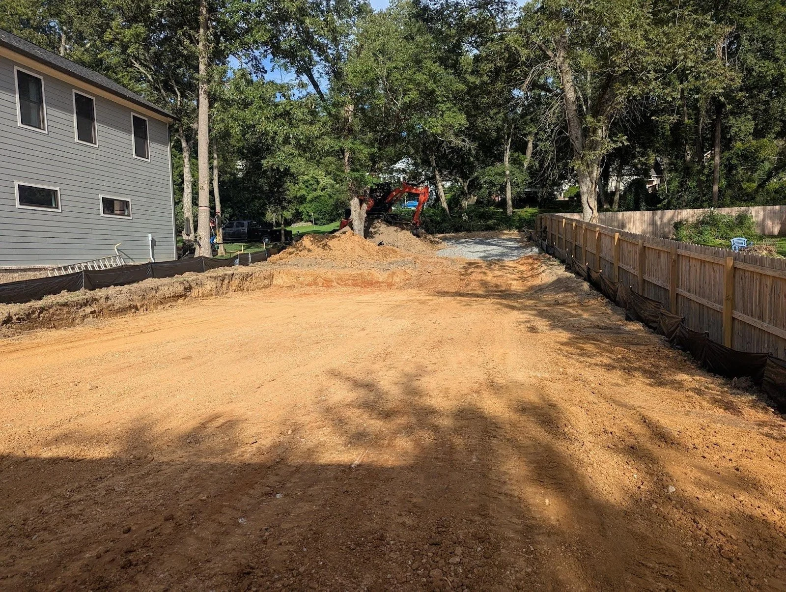 A backyard under construction with a dirt ground, a wooden fence on the right, and a construction vehicle near a tree in the background.
