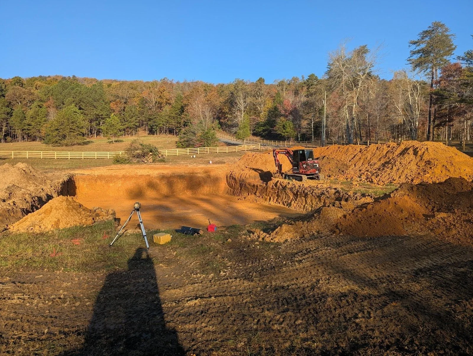 A construction site with a deep excavation pit, a mini excavator working on the side, and surveying equipment on the ground. Piles of dirt surround the area, with trees and a fence in the background under a clear blue sky.