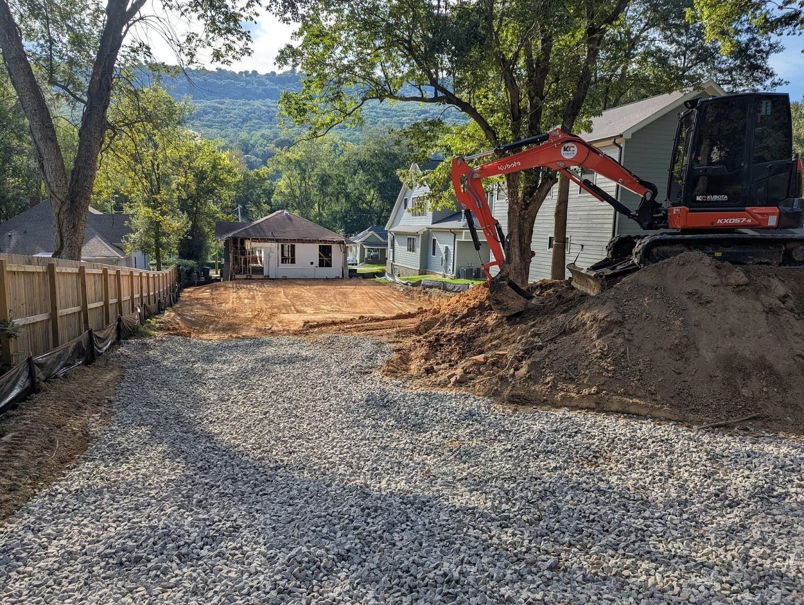 A construction site shows a gravel pathway leading to a cleared dirt area with an excavator digging. The background features trees, neighboring houses, and a shaded gazebo.