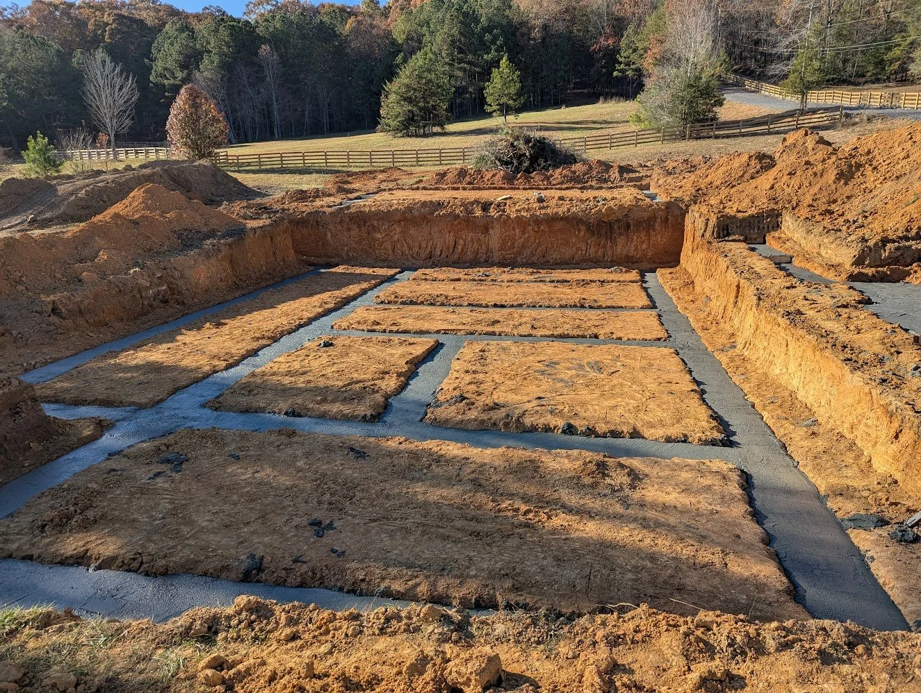 Excavation site with rectangular foundation trenches filled with concrete, surrounded by dirt, with trees and a fence in the background.