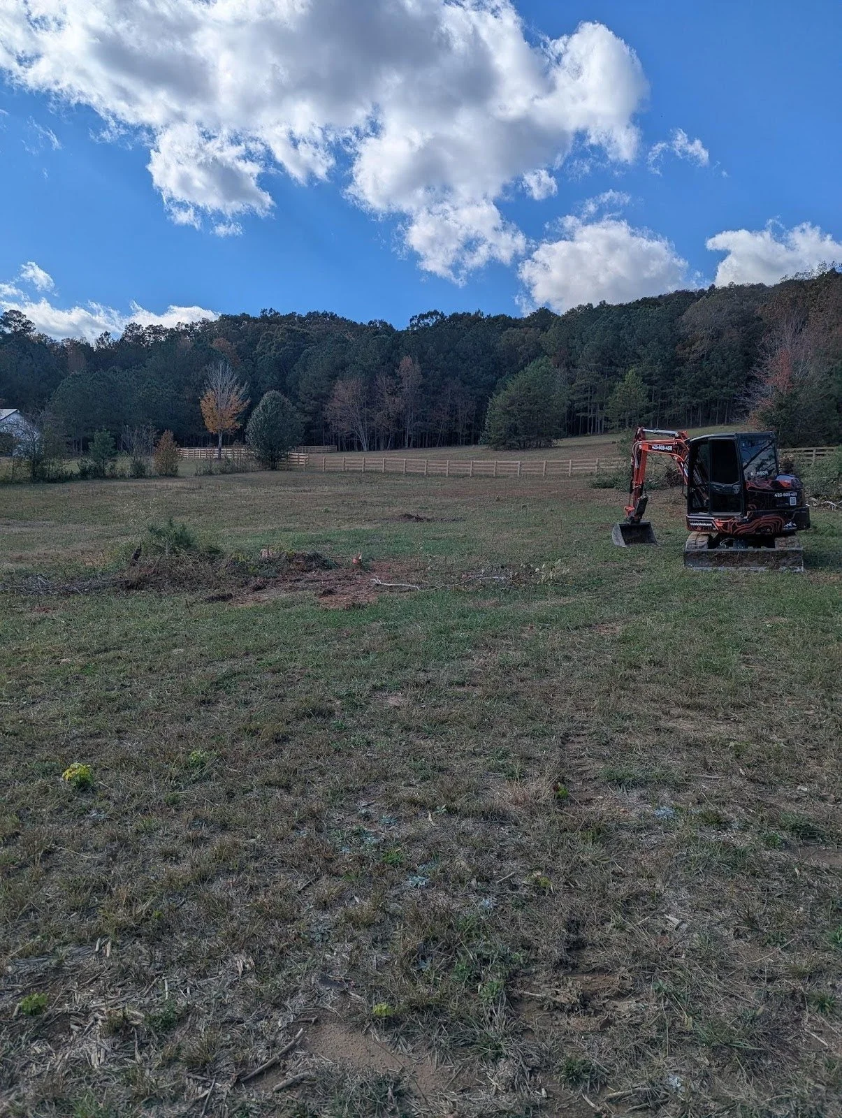A cleared grassy field with a small red and black mini excavator on the right, surrounded by trees and a wooden fence in the background, under a blue sky with white clouds.
