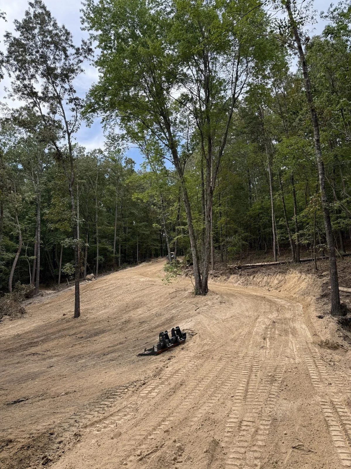 A dirt trail winding through a wooded forest with green trees, tire tracks in the dirt, and a small black tracked machine or equipment on the ground.