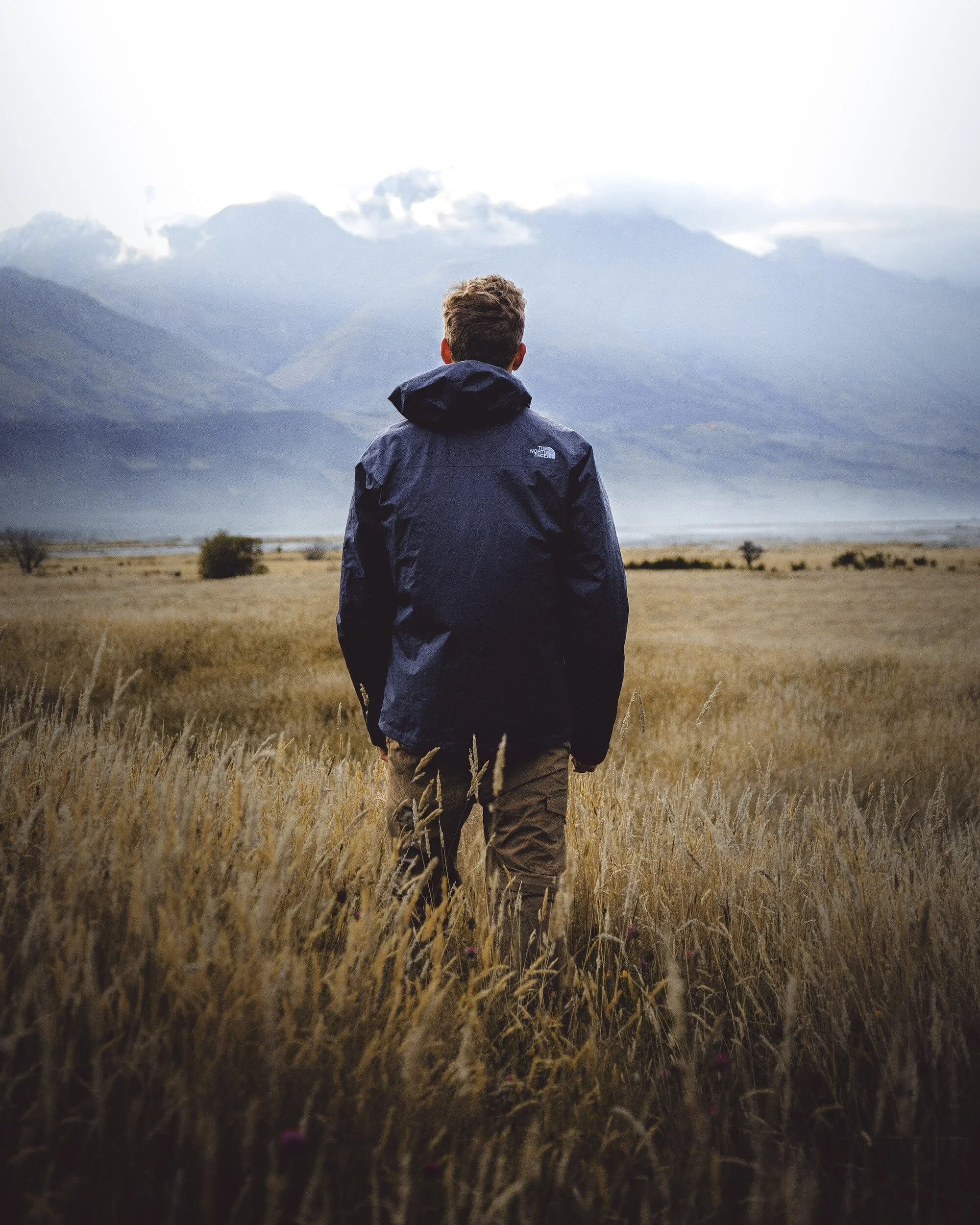 Person in blue jacket standing in a field facing mountains with clouds.