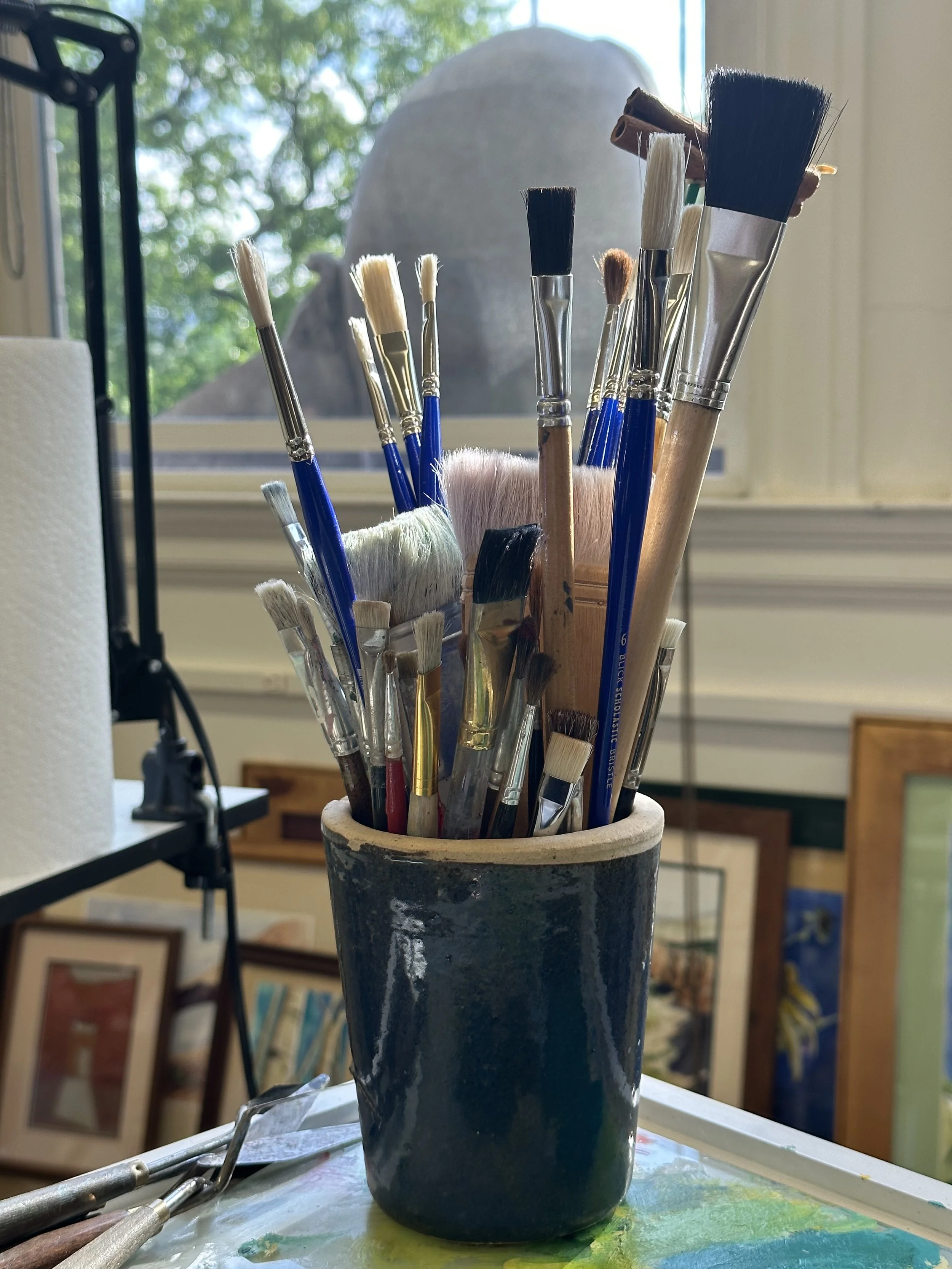 Assorted paintbrushes in a ceramic holder near a window with natural light, in an art studio setting.