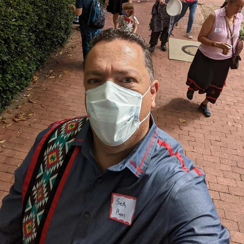 A man wearing a face mask and a name tag that reads 'Seth Mays' taking a selfie outdoors on a brick sidewalk with several people in the background.