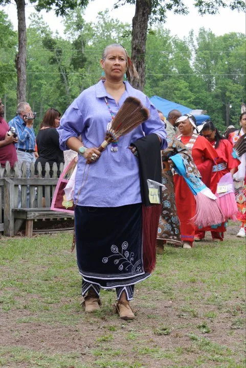 Woman in traditional clothing holding a broom at outdoor cultural event with other people in traditional attire in the background.