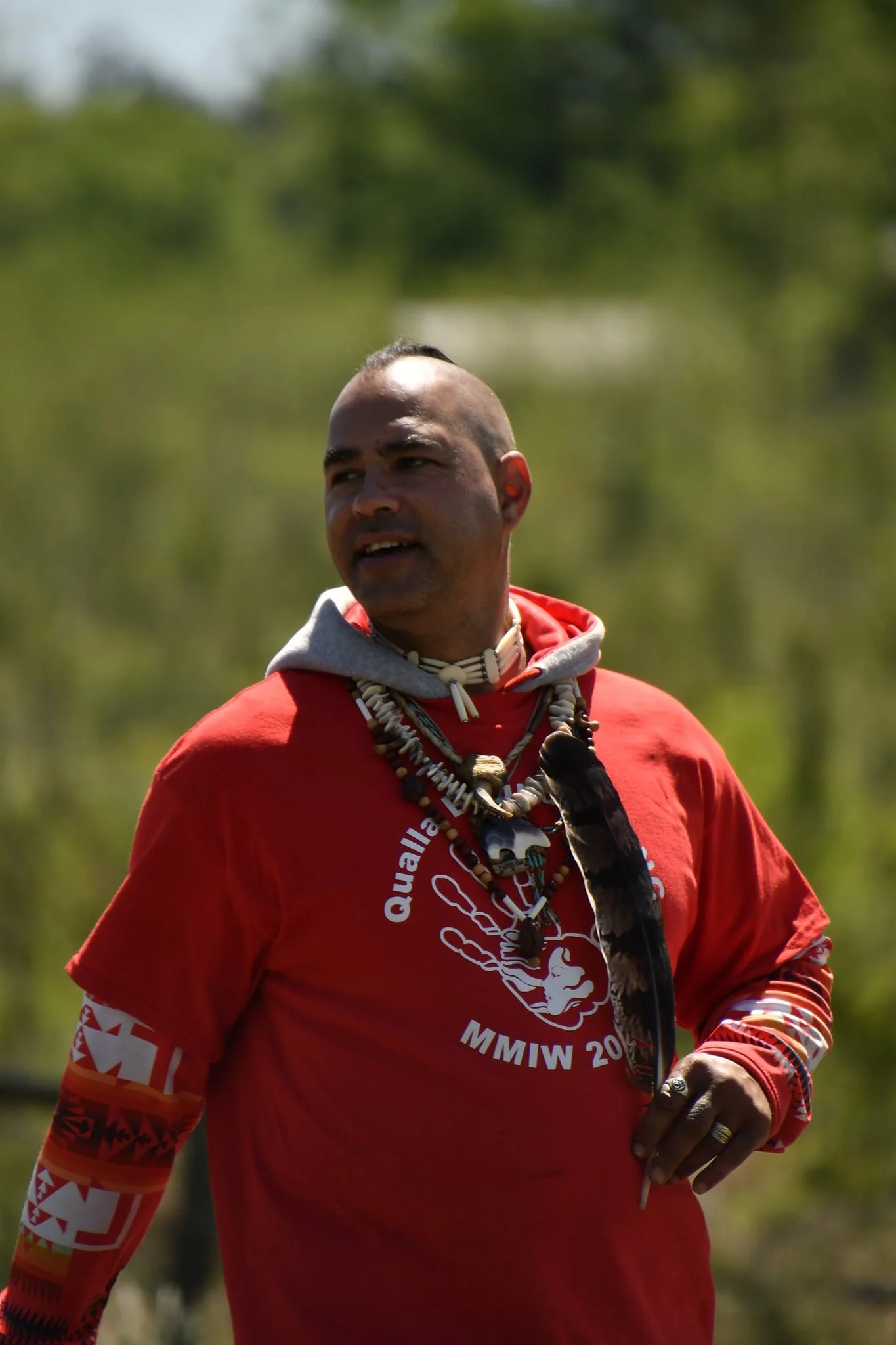 A man with a mohawk hairstyle wearing a red shirt and multiple necklaces, standing outdoors with greenery in the background.