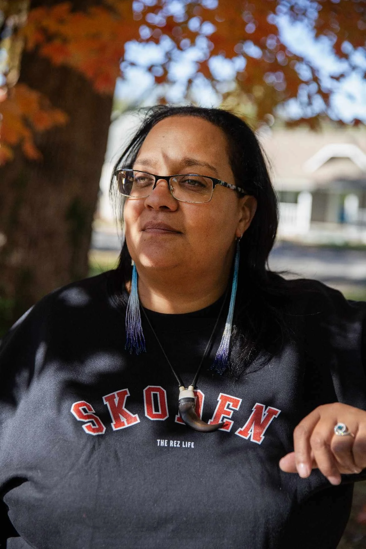A woman with glasses and long earrings is sitting outdoors under a tree with red and orange leaves. She is wearing a black sweatshirt with red and white text and a necklace with a large dark bead. She has long dark hair and is smiling slightly while looking off to the side.