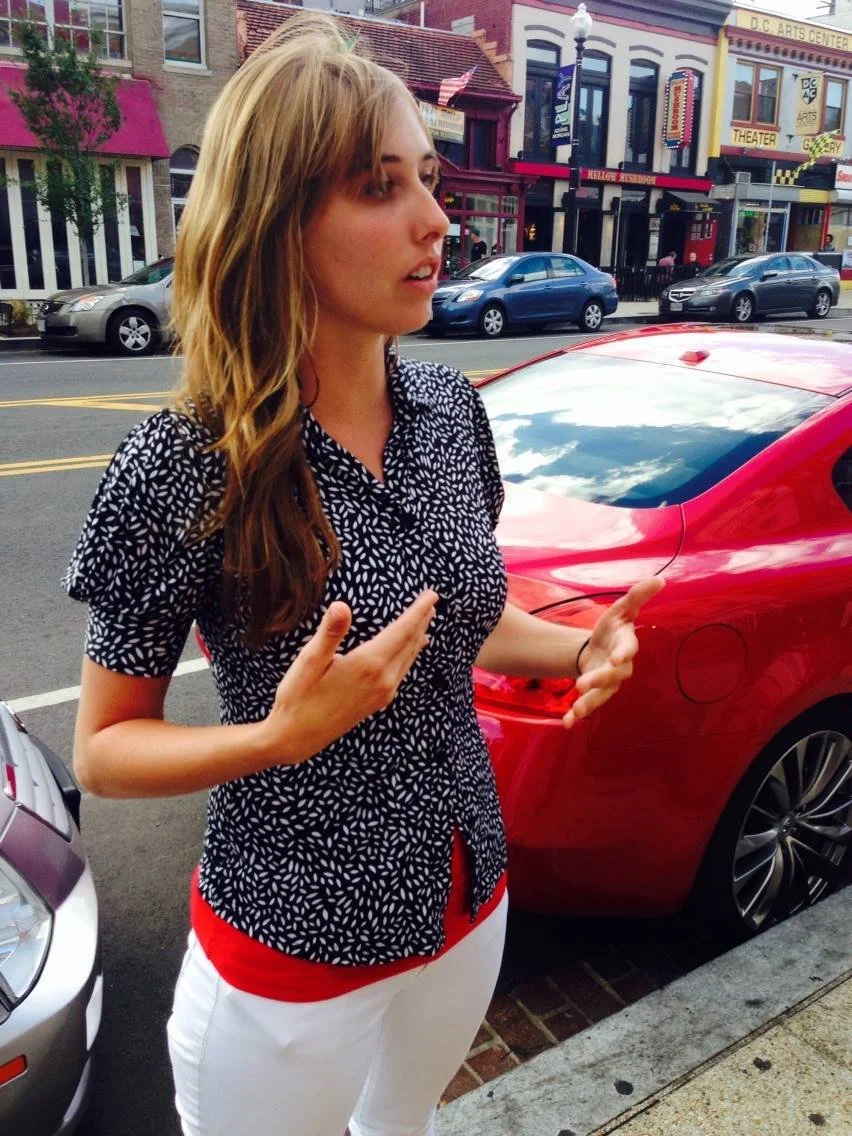 A woman standing on a city sidewalk near parked cars, gesturing with her hands, wearing a black and white patterned shirt and white pants with a red waistband, with colorful storefronts in the background.