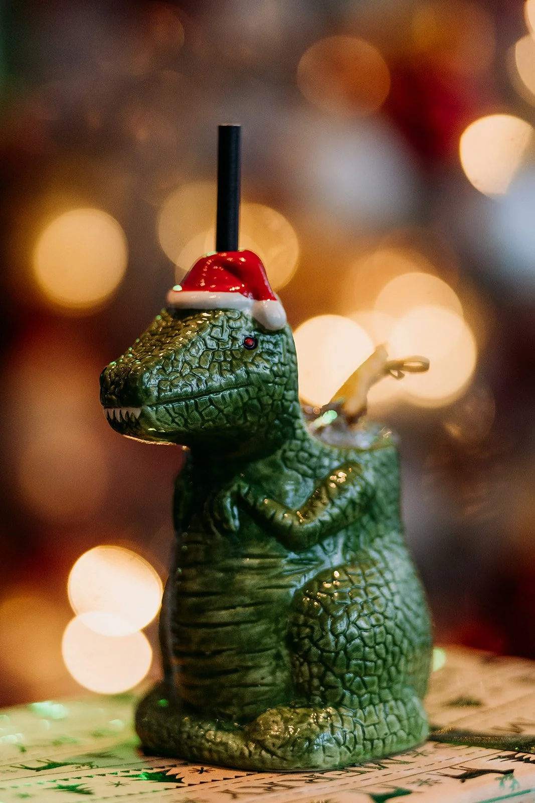 Dinosaur-shaped Christmas ornament wearing a Santa hat with a straw in its head, set against a blurred background of warm holiday lights at Miracle Pop Up Bar at The Raven Room in Whistler BC Canada.