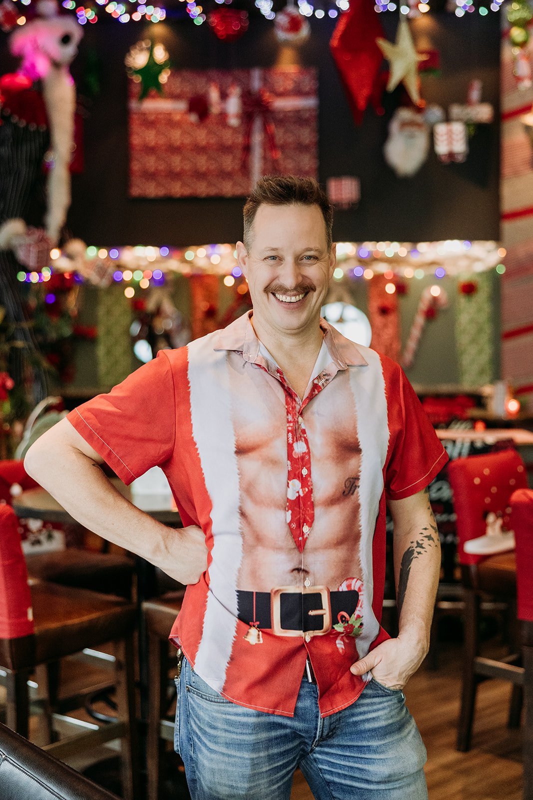 Luke Woodnutt smiling with a mustache, wearing a Christmas-themed shirt resembling Santa's suit with a printed muscular chest, standing inside a festively decorated room with Christmas ornaments, lights, and presents at The Raven Room in Whistler BC