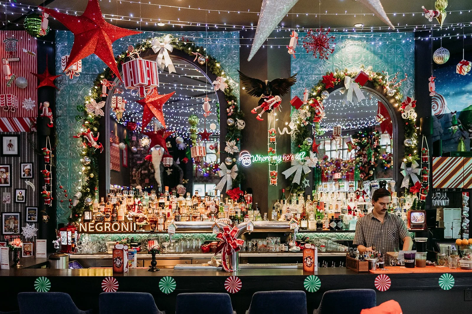 A festive bar decorated with Christmas ornaments, lights, and holiday-themed decorations, featuring a bartender preparing drinks at Miracle Pop Up Bar at The Raven Room in Whistler BC Canada.