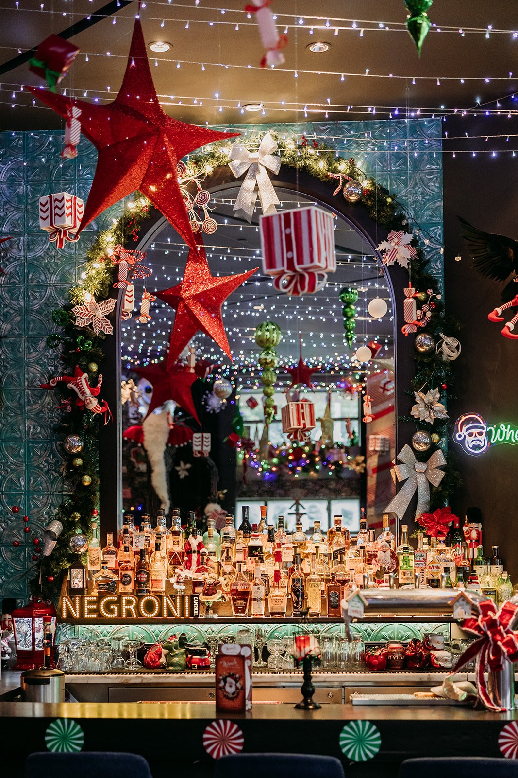 The Raven Room in Whistler decorated with Christmas ornaments, including large red stars, wrapped gift ornaments, and a green neon sign. The bar counter is filled with various bottles of spirits.