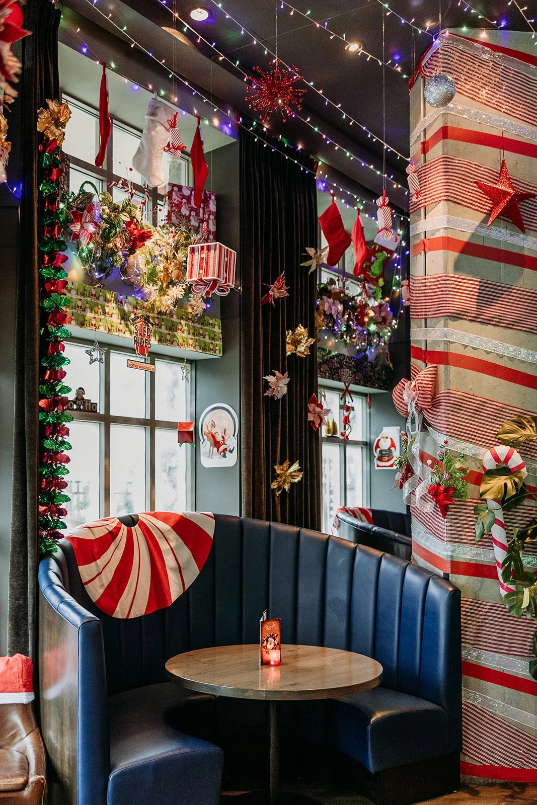 This image shows a cozy Christmas-themed cafe corner decorated with string lights, hanging stockings, wrapped gifts, and festive wreaths. There is a circular table with a candle and holiday decor at Miracle Pop Up Bar at The Raven Room in Whistler BC