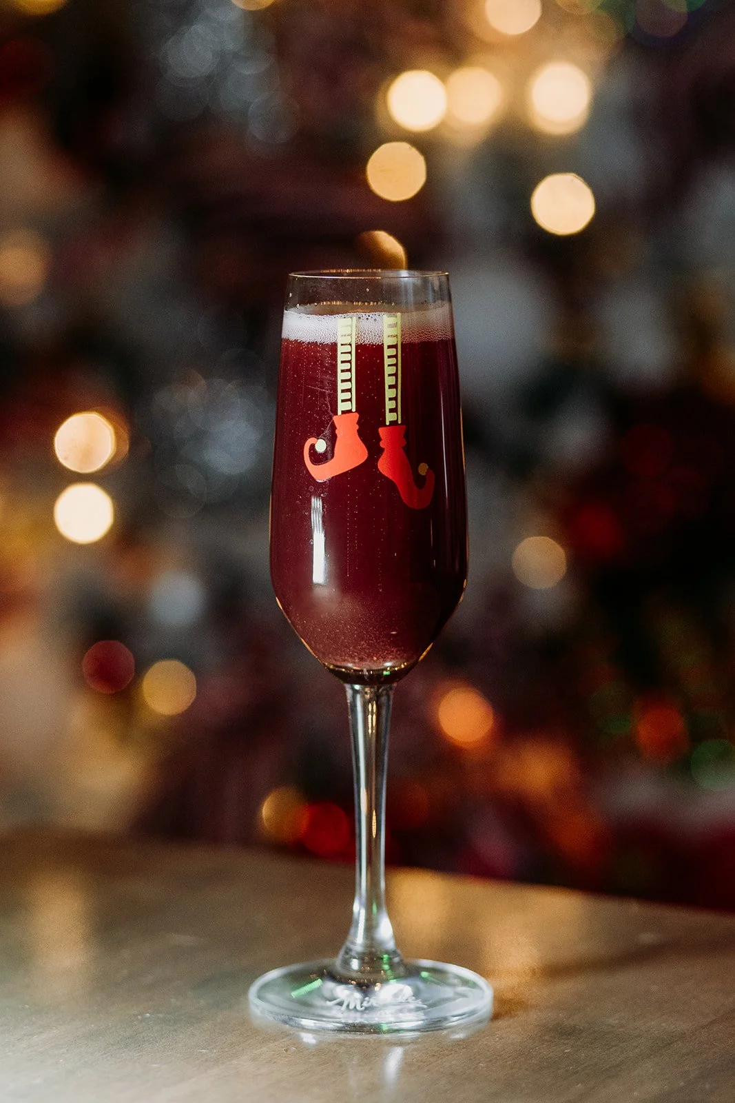 A glass of red sparkling beverage with two Christmas stocking decorations on the rim, with a blurred Christmas tree and lights in the background at Miracle Pop Up Bar at The Raven Room in Whistler BC Canada.