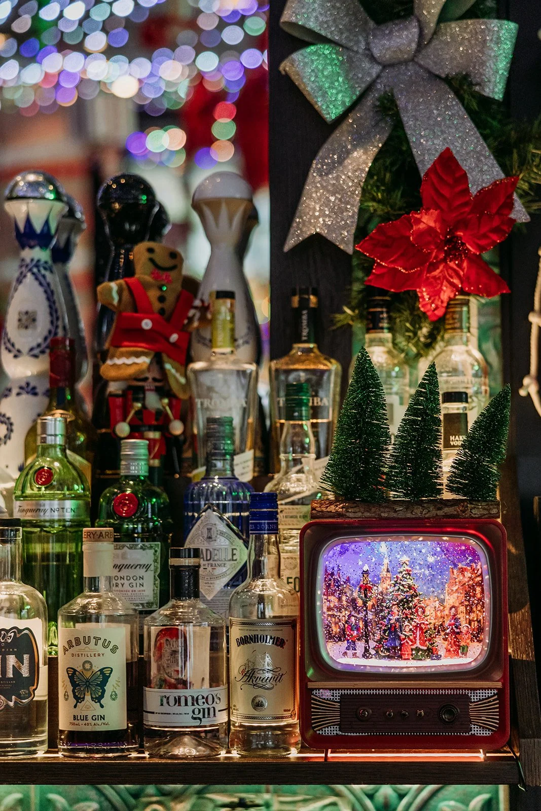 A holiday display with Christmas decorations, miniature Christmas trees, a small vintage TV showing a festive scene, various bottles of alcohol, a gingerbread man, and a poinsettia flower with silver ribbon in the background at Miracle Pop Up Bar