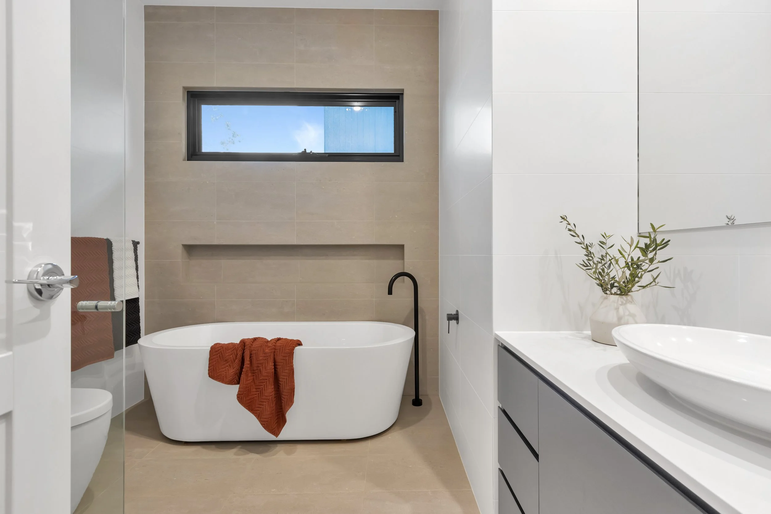 Modern bathroom remodel or renovation with bathtub, black faucet, gray vanity, white vessel sink, beige tile wall, white wall, window with blue sky, green plant in white vase, brown towel, and beige floor.