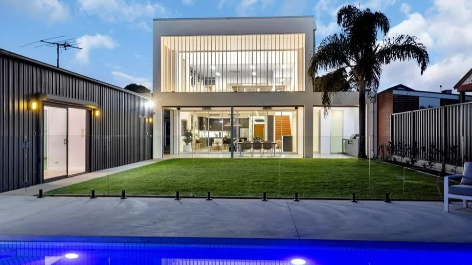 Modern house with large glass windows, a lawn, tall palm trees, and outdoor lighting, overlooking a swimming pool at sunset or dusk renovated by A Plus Property Services SA in Brooklyn Park Adelaide near Adelaide Airport