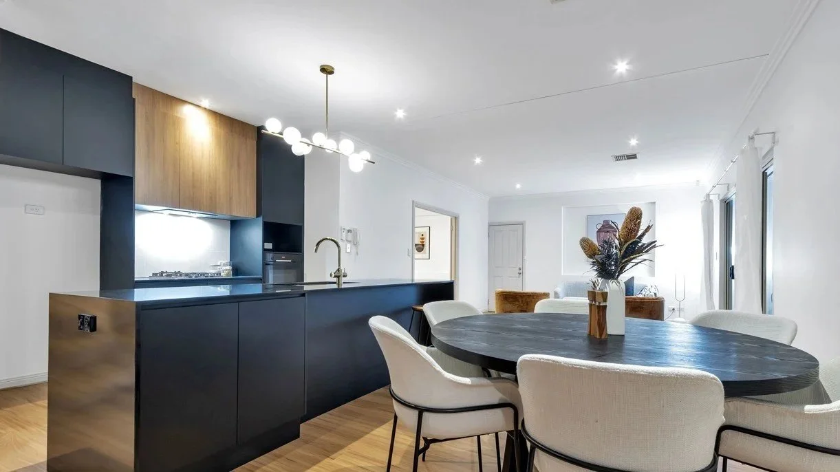 Modern open-plan living room and kitchen with black island, white chairs around a black dining table, and a bouquet of dried flowers in a vase in an Athelstone Adelaide residence renovated and painted by A Plus Property Services near Wadmore Park