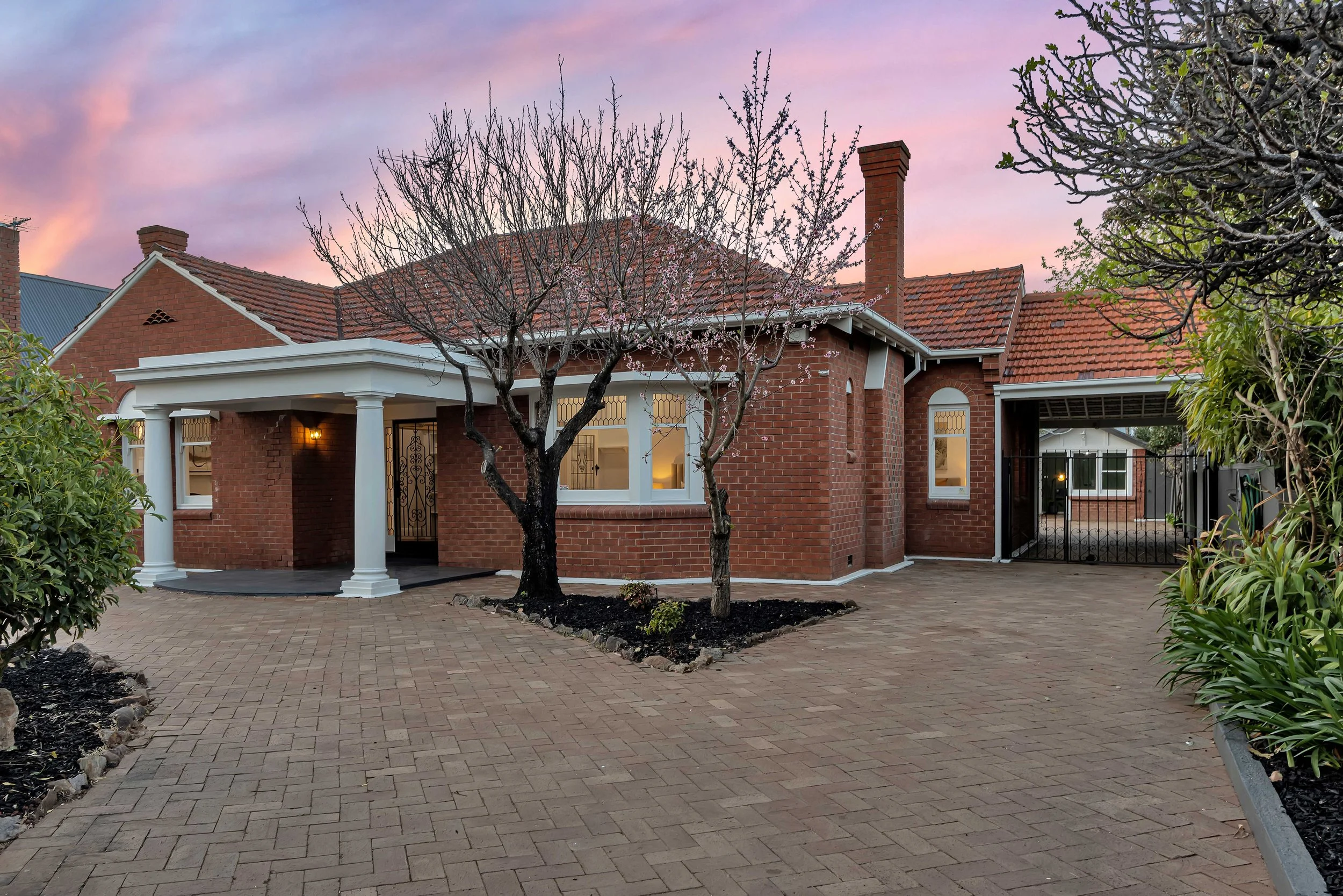 A brick house with a red tiled roof, a white front porch with columns, and a small tree in the front yard, photographed at sunset.