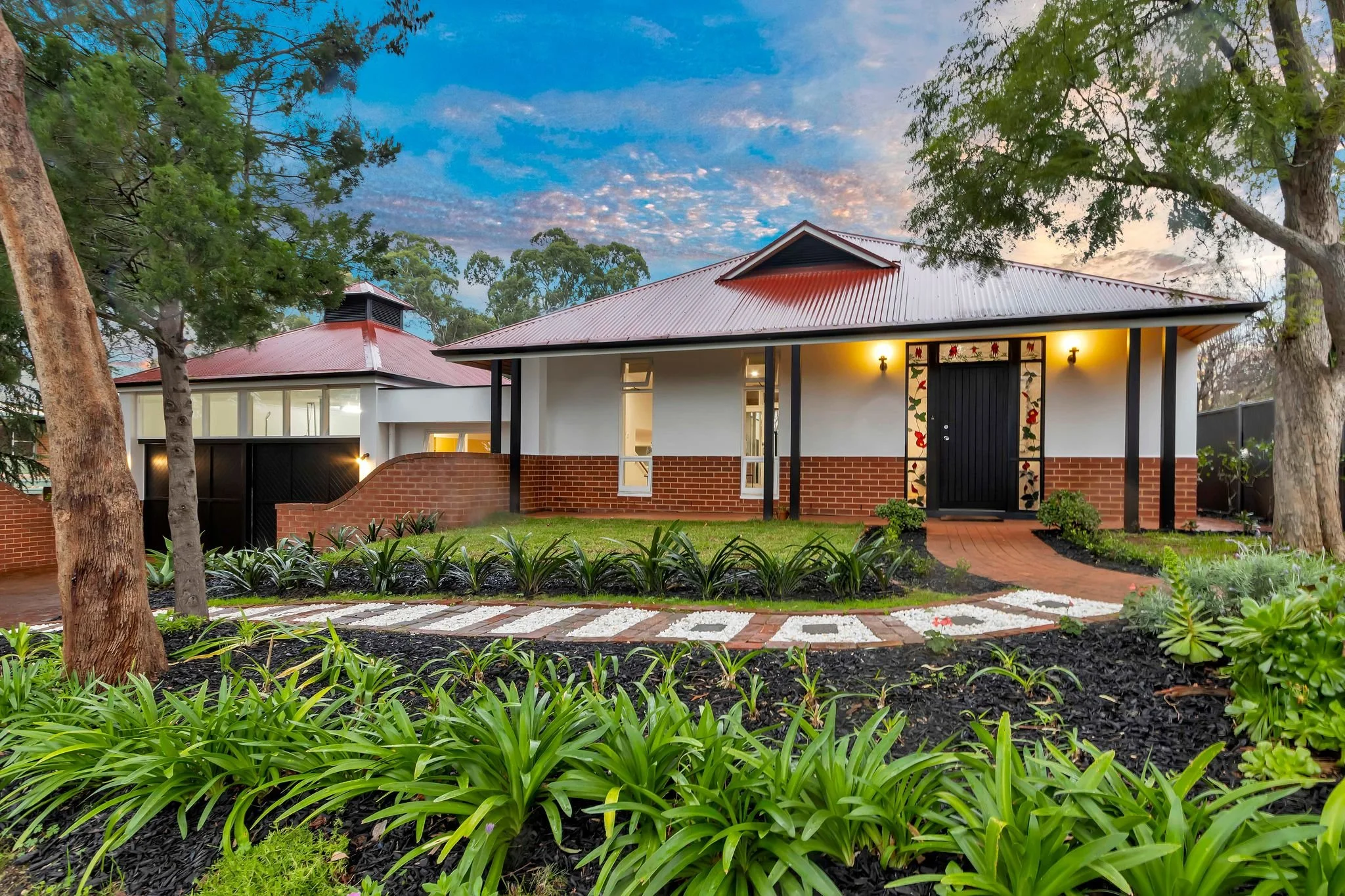 A  house with a red metal roof, white walls, and a brick base, surrounded by lush green plants and trees. The front door is black with decorative trim, and there are two small windows. The garden has a brick pathway leading to the entrance, with flower beds and landscaping lights.