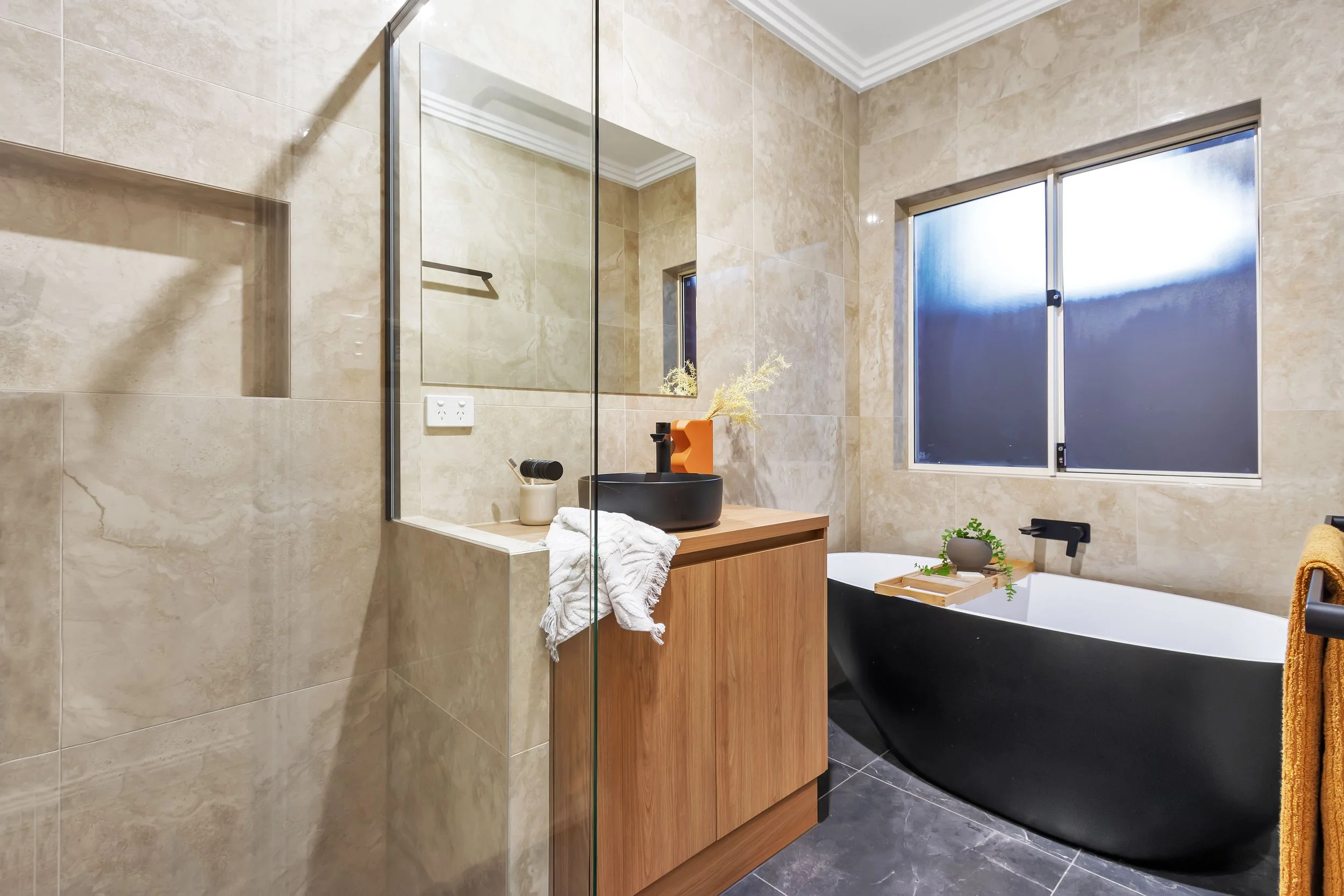 Modern bathroom remodel with beige tiled  in Adelaide walls, black freestanding bathtub, wooden vanity with black vessel sink, mirror, and window with frosted glass.
