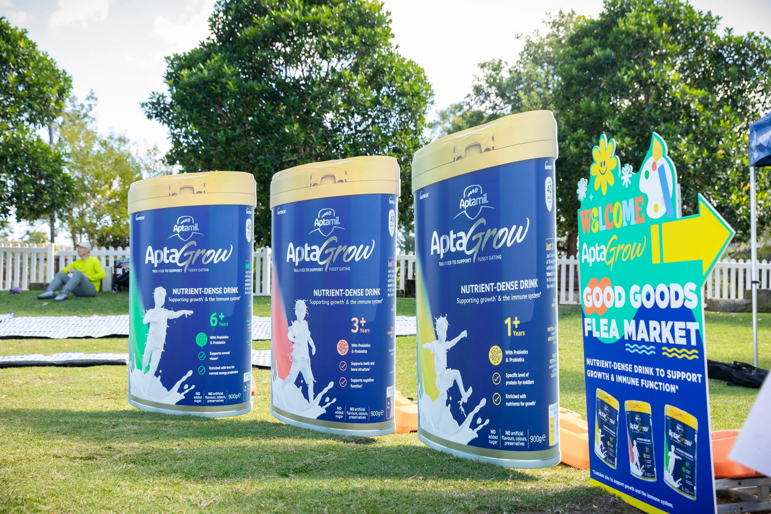 Outdoor display of large cans of Aptamil AptaGrow infant formula with colorful signage for a flea market, with a person sitting on a bench in the background.