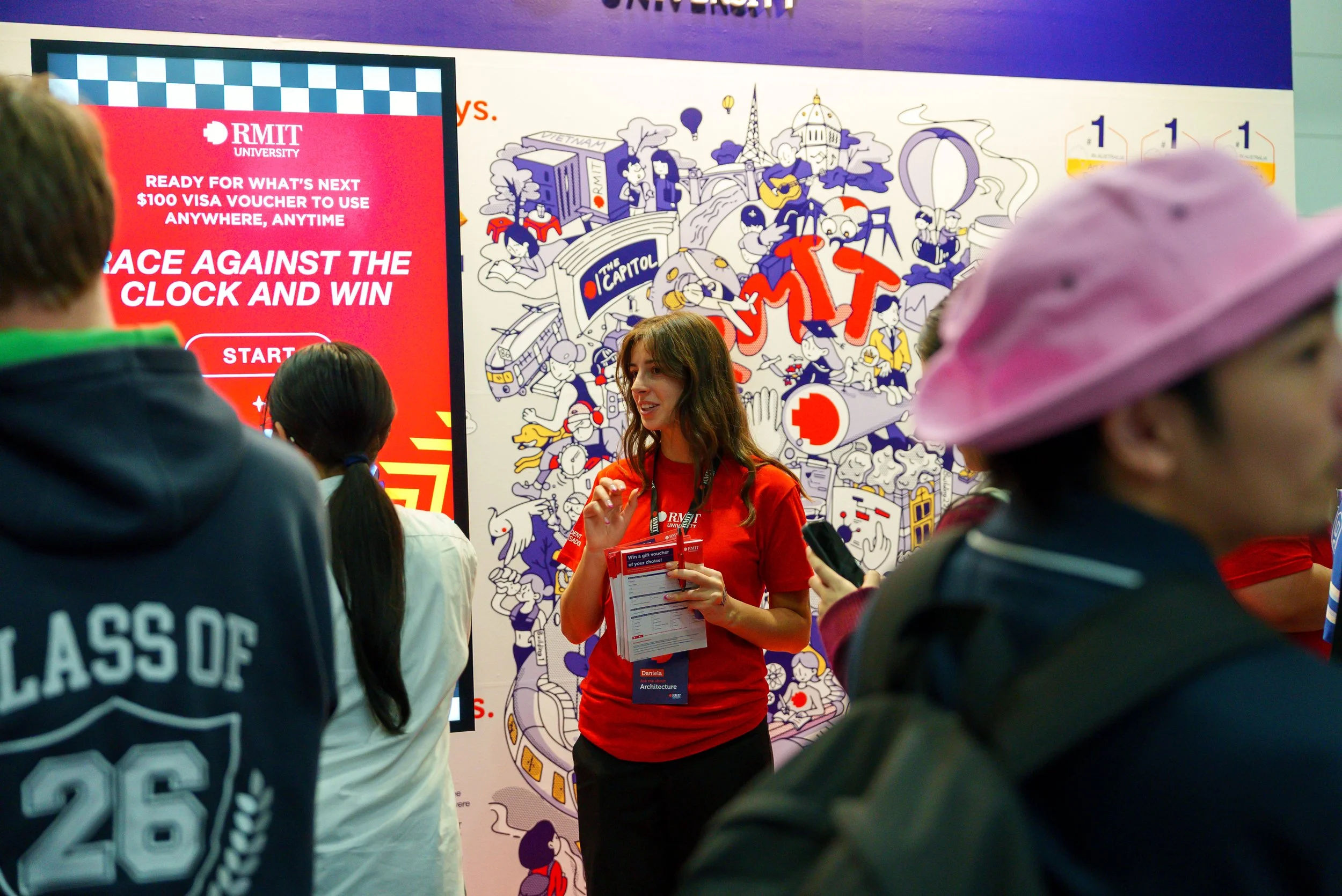 A woman with brown hair, wearing a red shirt and lanyard, is holding papers and talking to a group of people at an indoor event. Behind her, there is a colorful mural with cartoon-style illustrations of city landmarks, people, and animals. To the lef